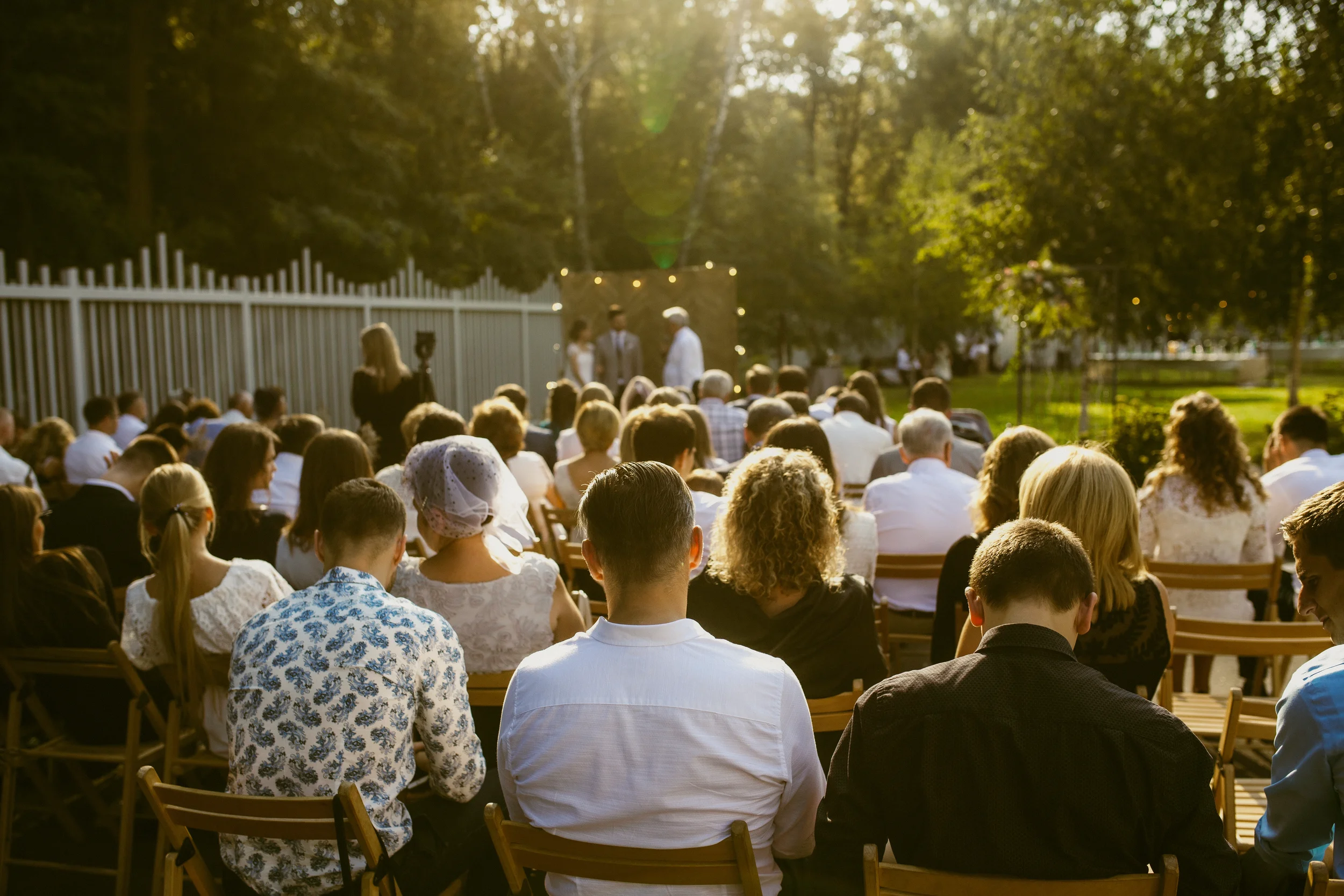 Menschen sitzen an Tischen im Freien und beobachten eine Hochzeit im Sonnenlicht, während im Hintergrund eine Hochzeitszeremonie stattfindet.