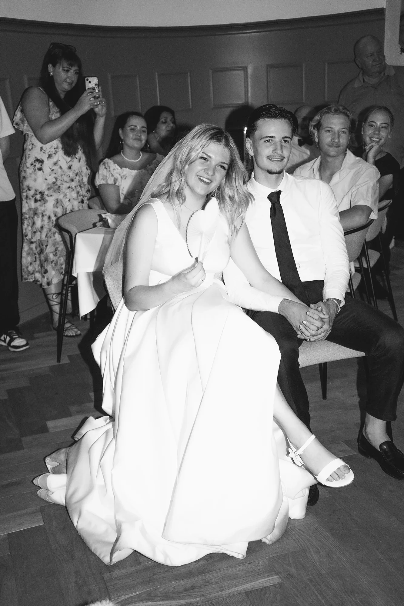 Eine Gruppe von Menschen bei einer Feier, im Vordergrund ein lachendes Brautpaar, die Frau trägt ein weißes Kleid und die Schuhe haben einen Absatz. Im Hintergrund stehen weitere Gäste, einige machen Fotos.