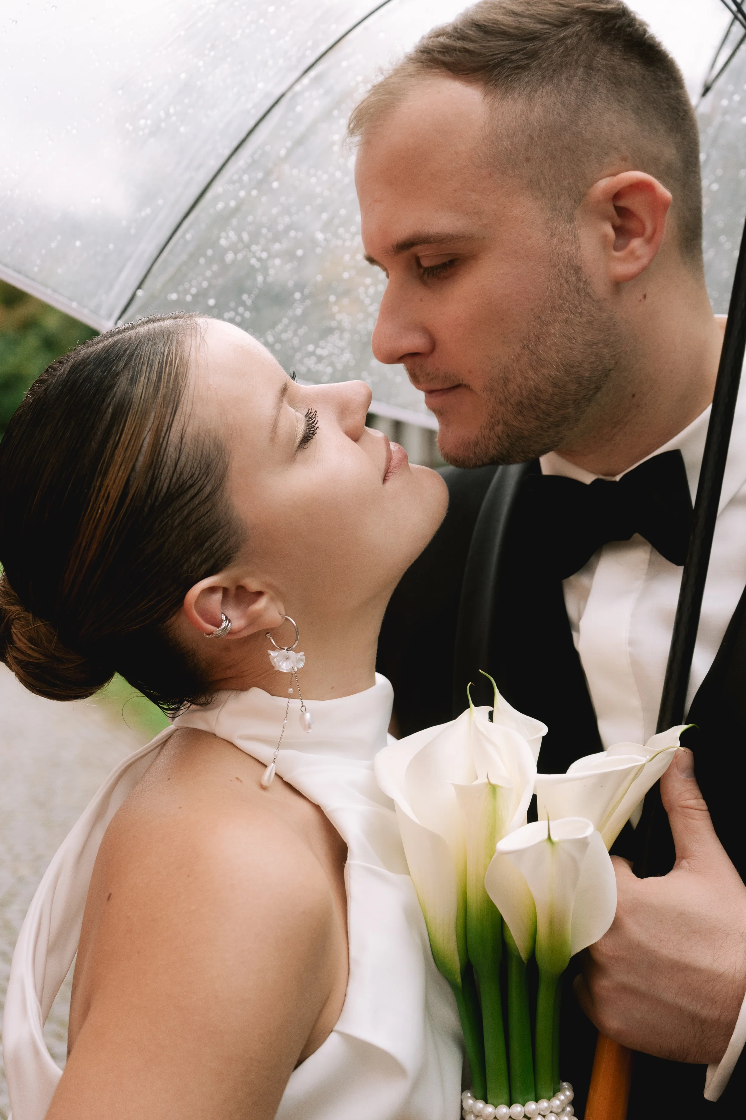 Ein Paar bei einer Hochzeitszeremonie, das sich vorsichtig berührt, mit einer Unterlage unter einem transparenten Regenschirm, der Regentropfen aufweist. Die Frau trägt eine elegante silberne Ohrringe und eine weiße Brautkleidung, während der Mann ei