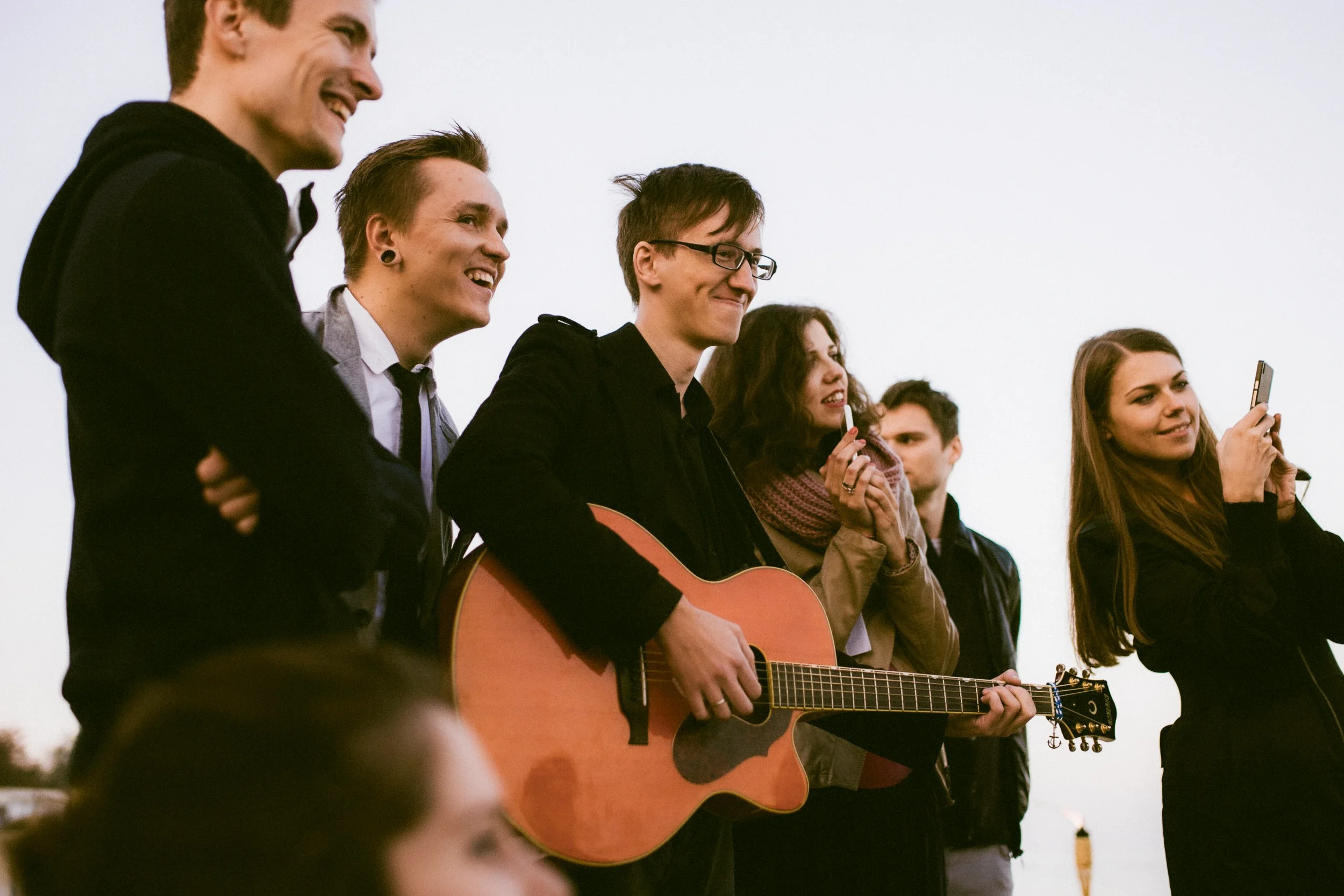 Eine Gruppe junger Menschen, die zusammen im Freien Musik macht, mit einer Person, die Gitarre spielt, während andere singen oder mit einem Telefon aufnehmen.