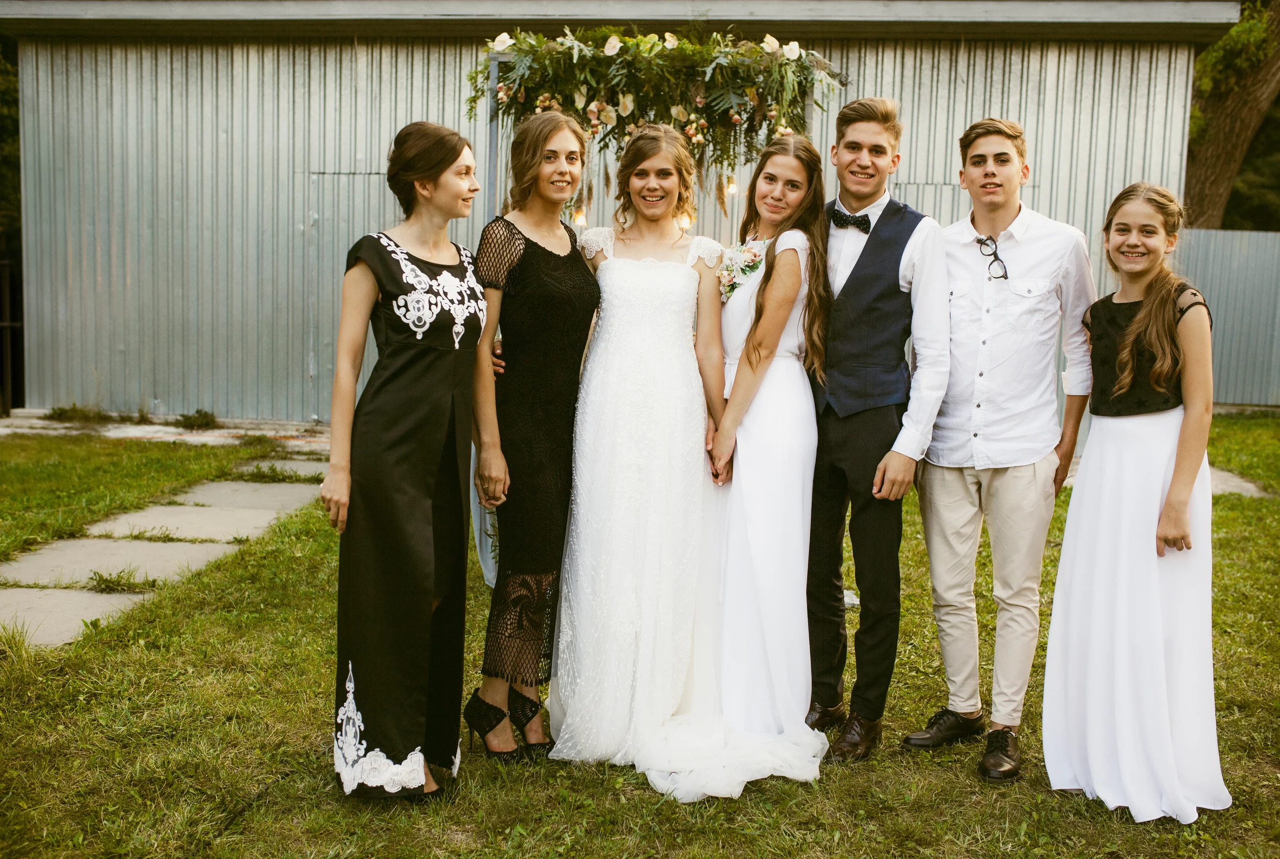 Eine Gruppe von sieben jungen Erwachsenen steht bei einem Hochzeitsempfang im Freien vor einer dekorativen Blumeninstallation.