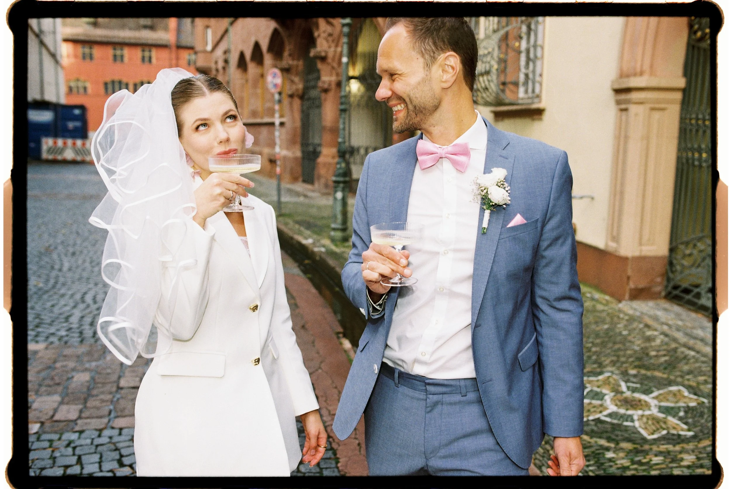 Ein Paar beim Hochzeitsempfang auf einer Kopfsteinpflasterstraße, der Bräutigam trägt einen blauen Anzug mit rosa Fliege, die Braut trägt ein weißes Kleid mit Schleier und hält ein Champagnerglas, beide lachen sich an.
