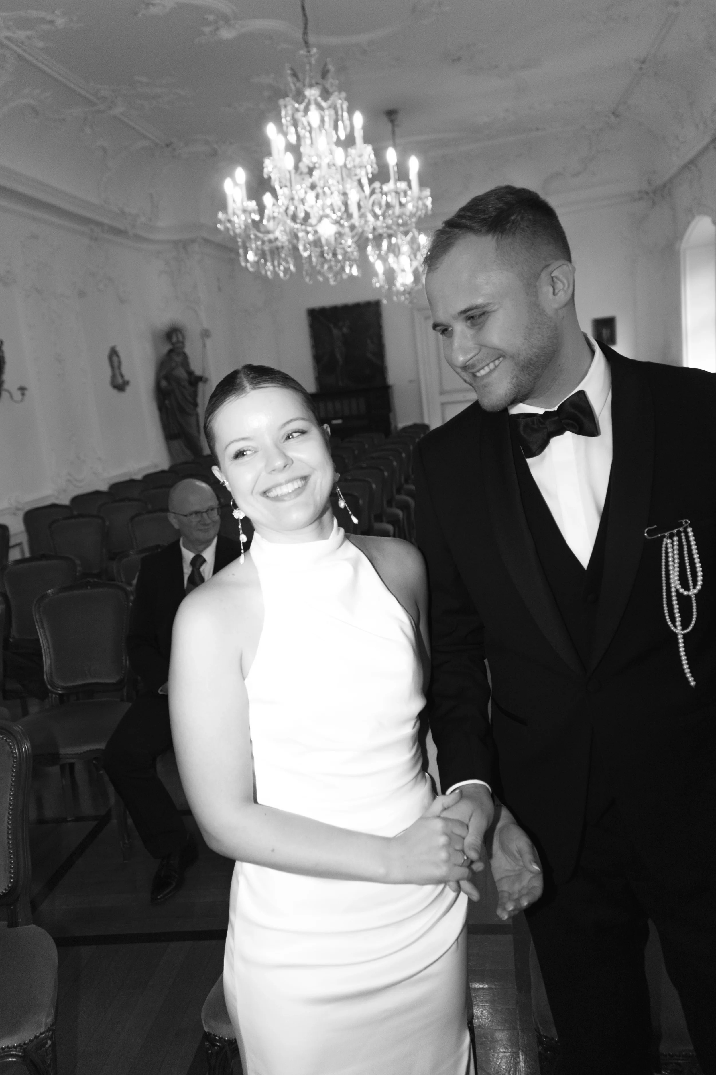 Ein glückliches Brautpaar bei einer Hochzeit, die sich die Hand geben, im eleganten Raum mit Kristallleuchter und barocken Wanddekorationen.