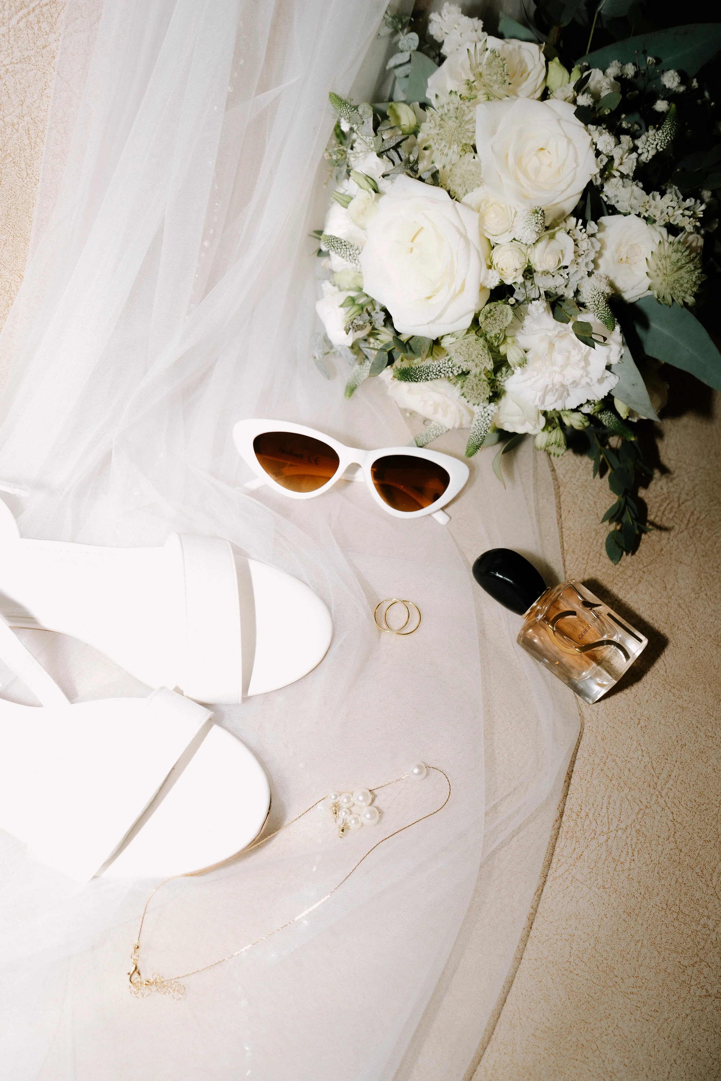 Weißer Brautstrauß mit weißen Rosen und grünem Laub, Sonnenbrille, Perlenkette, goldene Ringe, Parfümflasche, weißer Stuhl, weißes Tischtuch, weißer Schleier