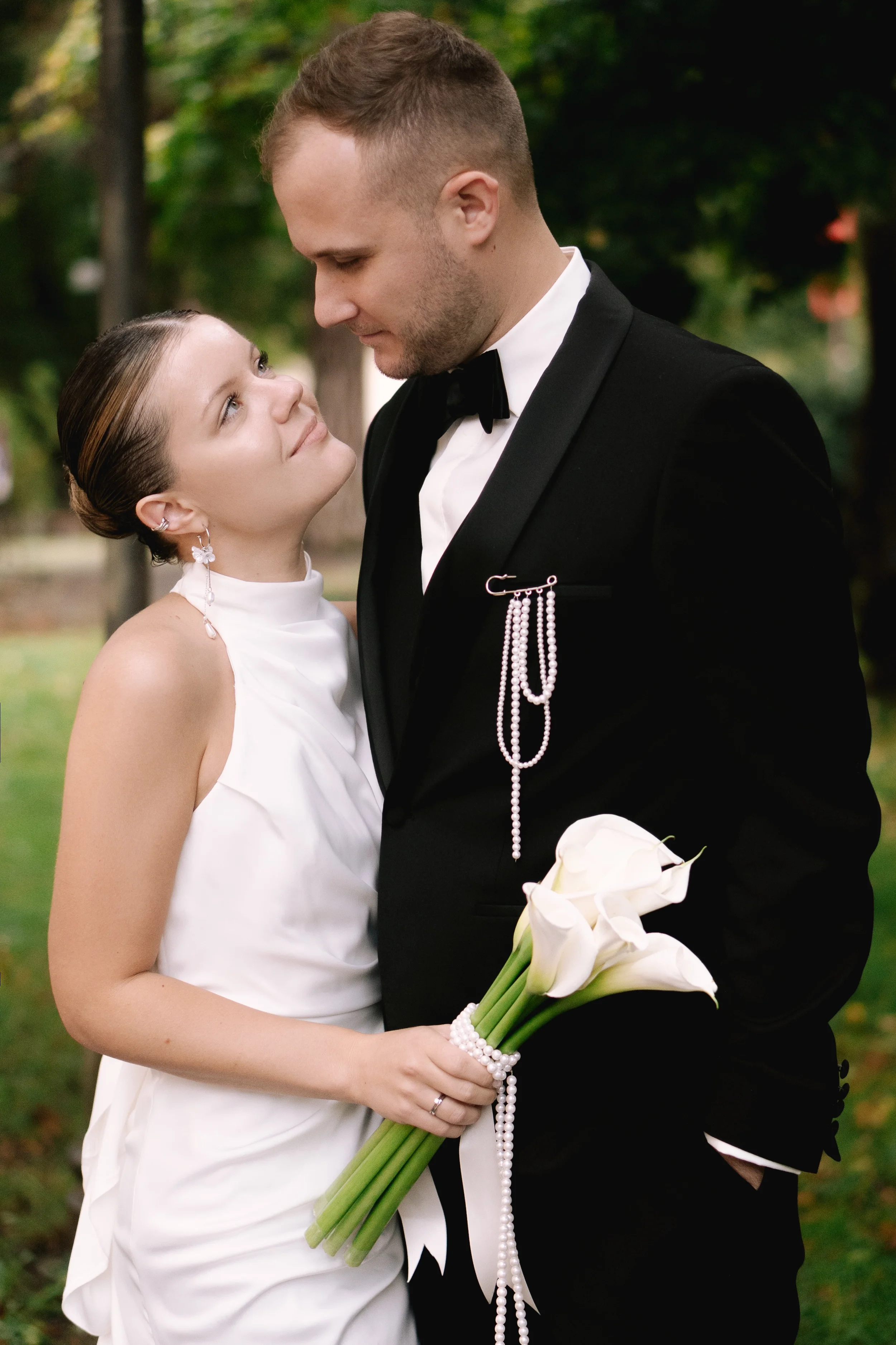 Ein Brautpaar bei der Hochzeit im Freien, die Braut hält einen Blumenstrauß aus weißen Calla-Lilien, beide schauen sich liebevoll an.