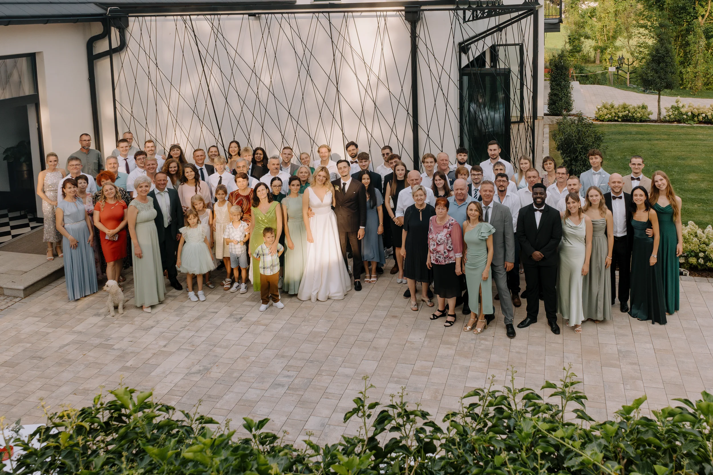Eine große Gruppe von Menschen, darunter Erwachsene und Kinder, posiert vor einem modernen Gebäude mit geometrischem Wandmuster. Es ist wahrscheinlich eine Hochzeit oder Familienfeier im Freien.
