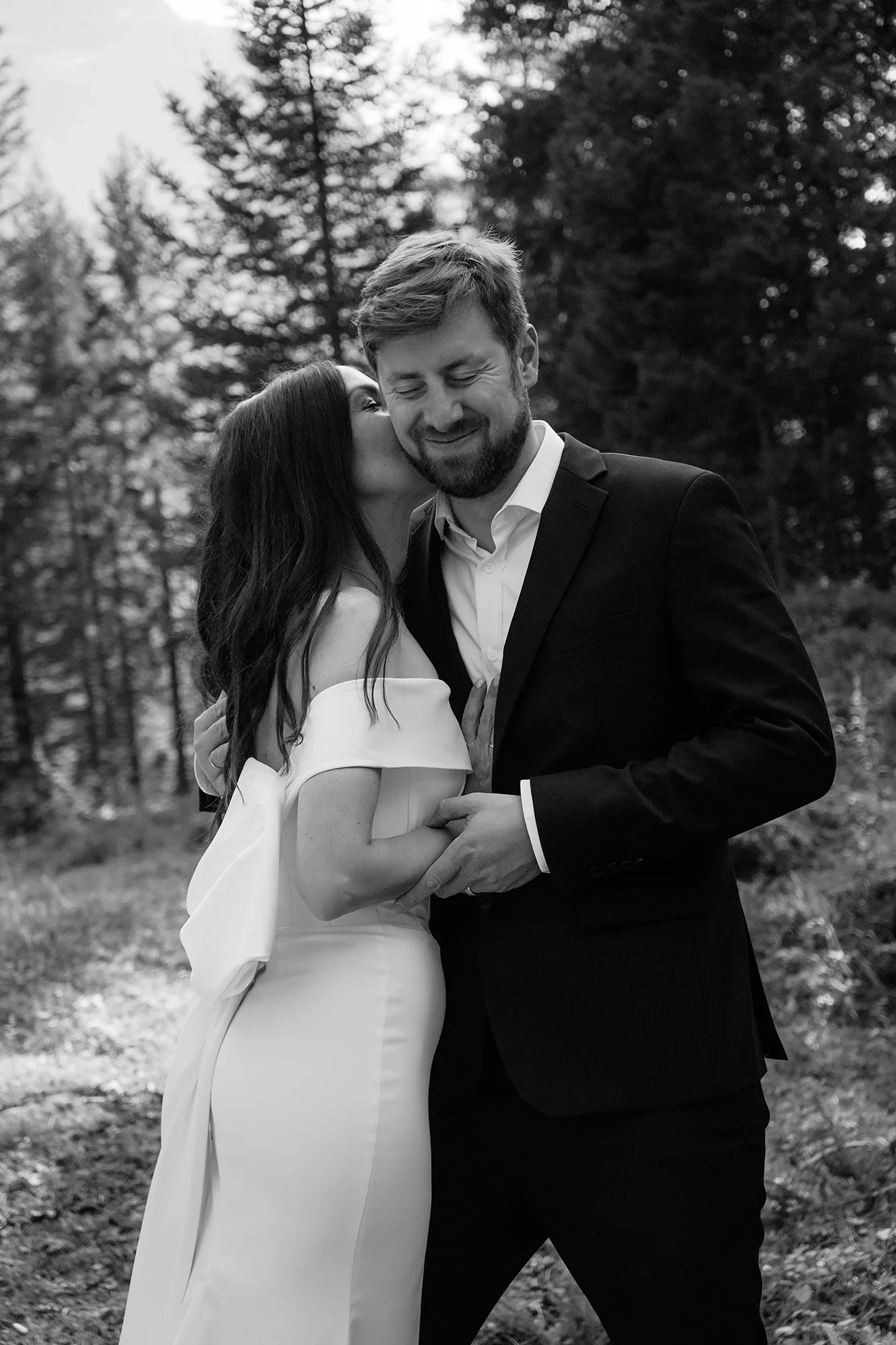 Schwarz-Weiß-Foto eines Liebespaares bei einer Umarmung im Freien, die Frau küsst den Mann auf die Wange, beide tragen formelle Kleidung, umgeben von Bäumen.