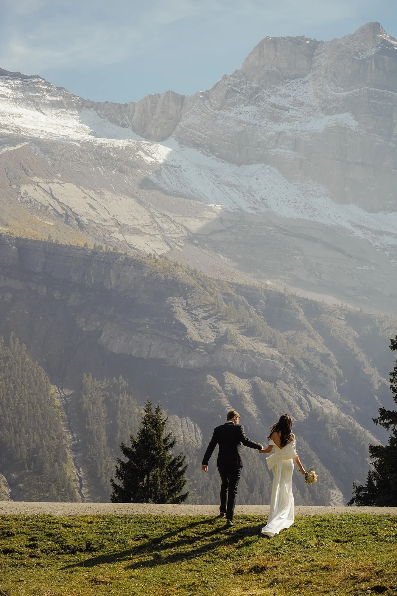 Ein Brautpaar hält sich an den Händen und läuft in einer malerischen Berglandschaft, umgeben von hohen Bergen mit Schnee und Tannenbäumen im Vordergrund.