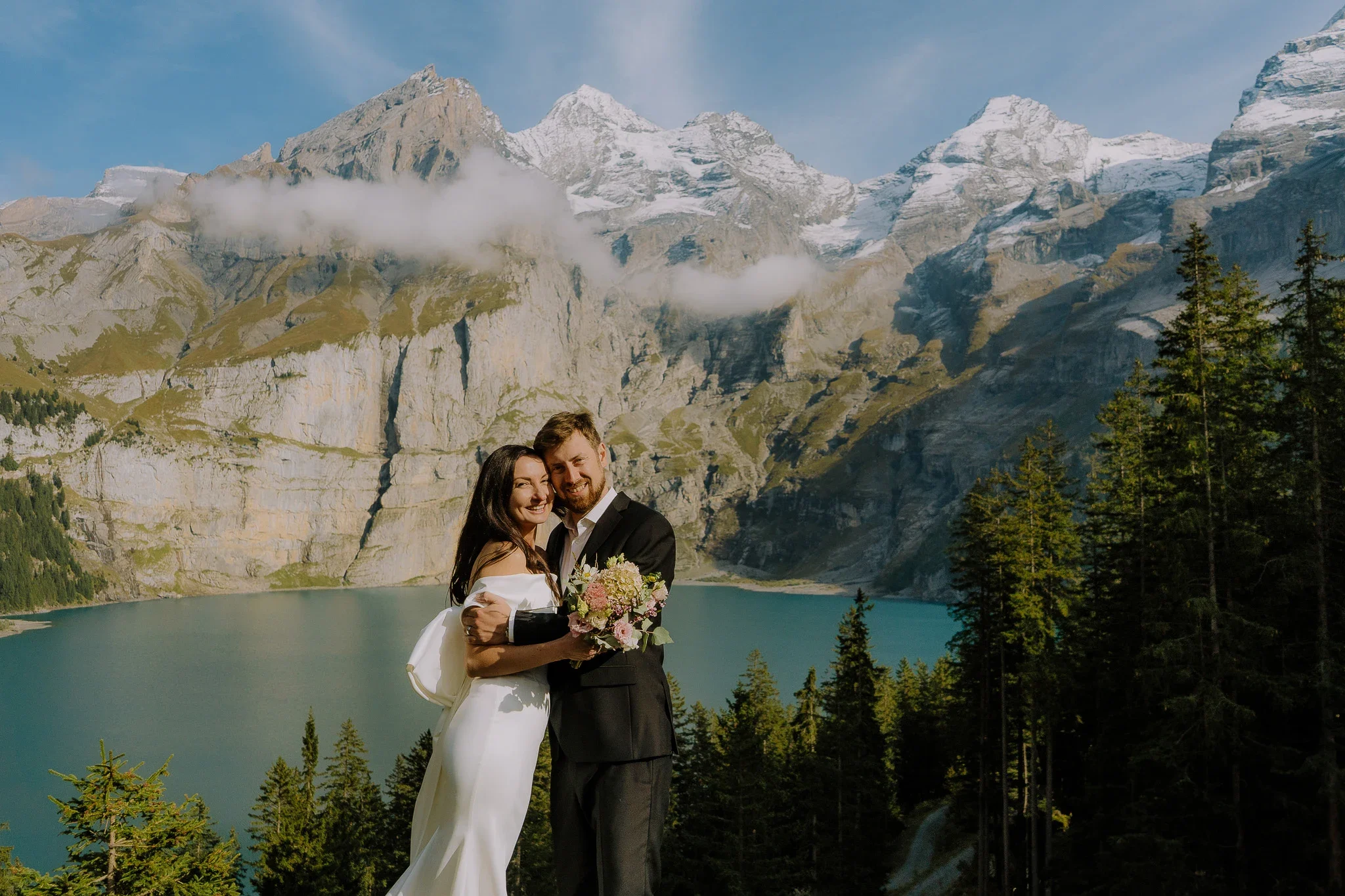 Ein verliebtes Paar in formeller Kleidung steht vor einer Bergkulisse mit einem See, umgeben von Tannenbäumen, in den Alpen.
