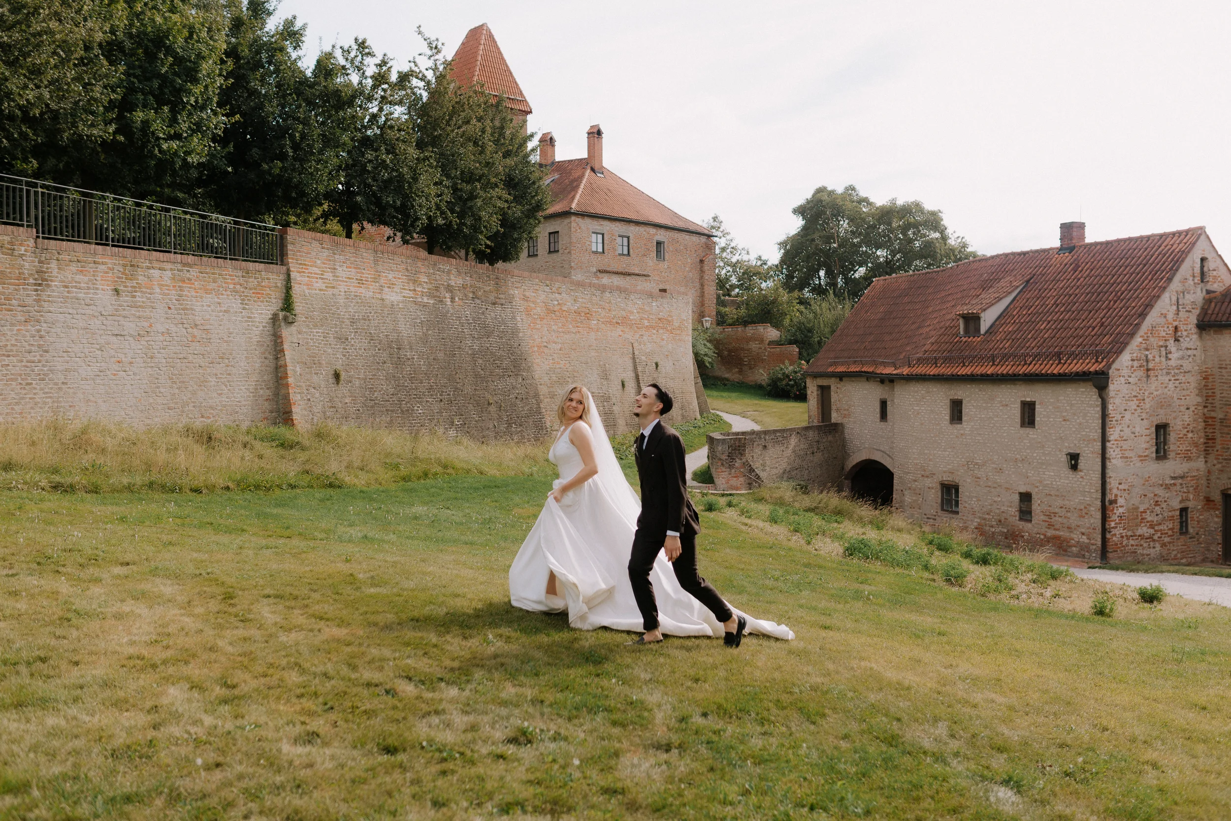 Ein Brautpaar in formeller Kleidung beim Spazieren auf einer Wiese vor einer alten Schlossmauer und historischen Gebäuden, umgeben von Bäumen.
