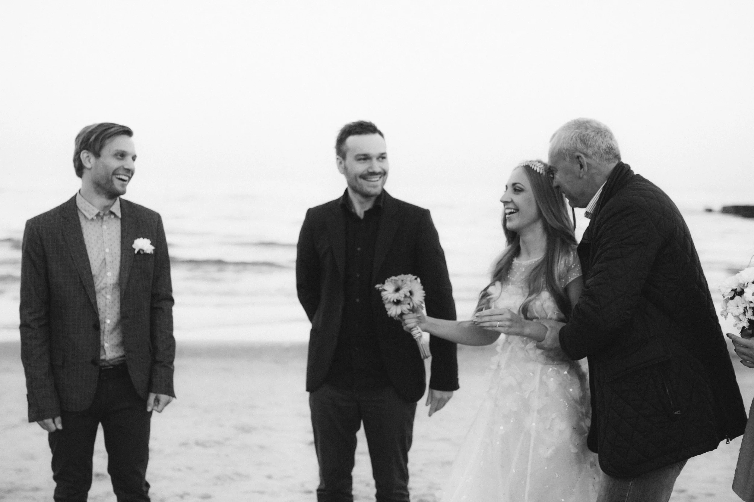 Vier Menschen bei einer Hochzeitszeremonie am Strand, happy und lachend, eine Frau mit Brautkleid und Blumenkranz auf dem Kopf, umgeben von drei Männern in formeller Kleidung.