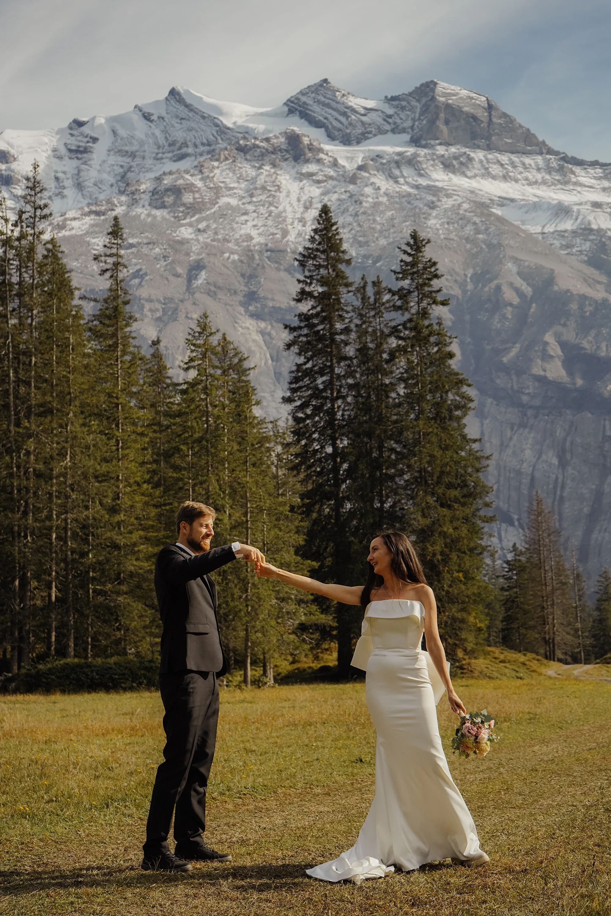 Ein Mann in einem schwarzen Anzug und eine Frau im weißen Brautkleid bei einer Hochzeitszeremonie im Freien vor einer Bergkulisse mit schneebedeckten Gipfeln. Der Mann reicht der Frau die Hand, während diese ein Blumenbouquet hält.
