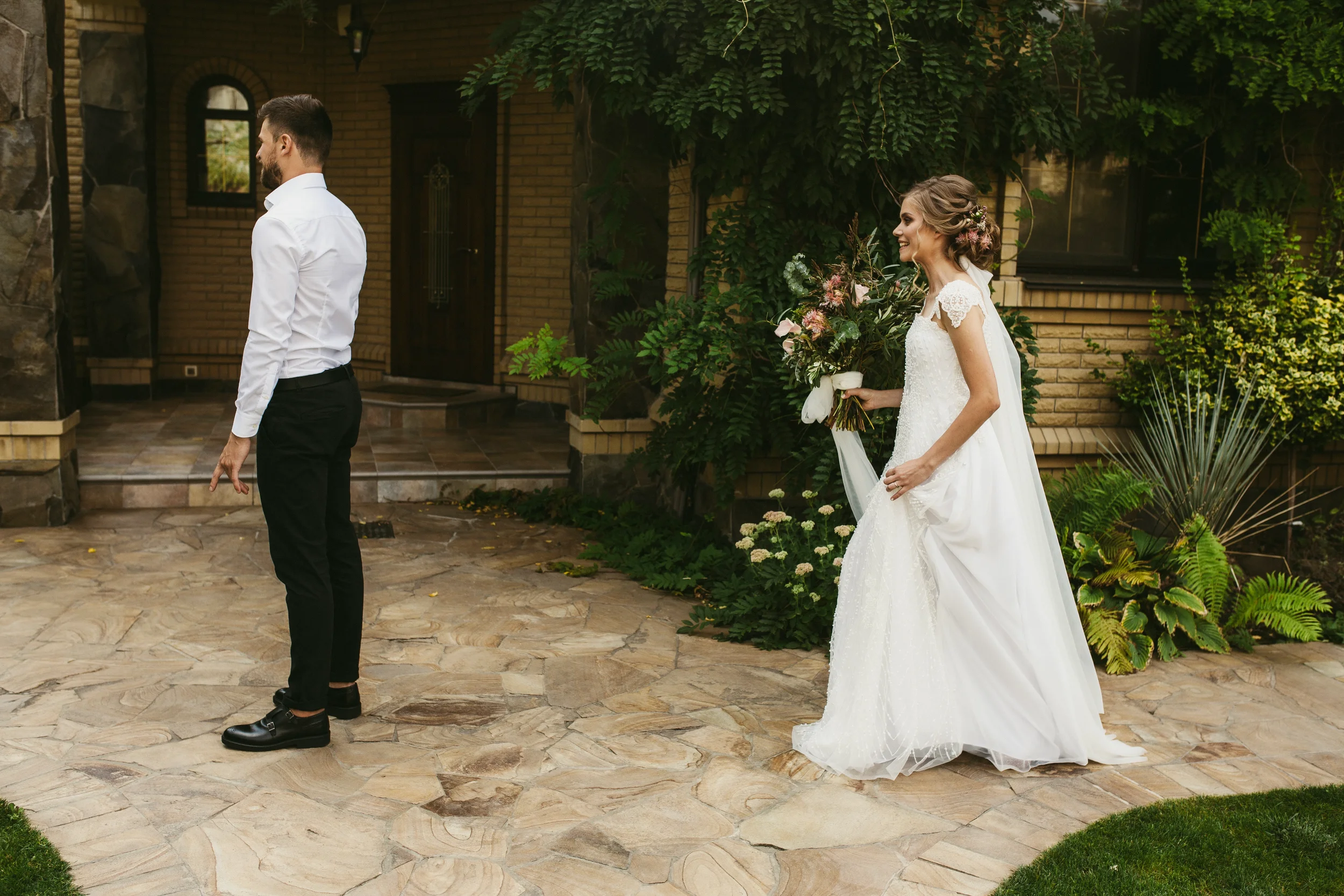 Braut in weißem Hochzeitskleid mit Blumen im Haar, hält einen Blumenstrauß, steht vor einem Mann in weißem Hemd und schwarzen Hosen, im Garten vor einem Haus mit viel Grün.