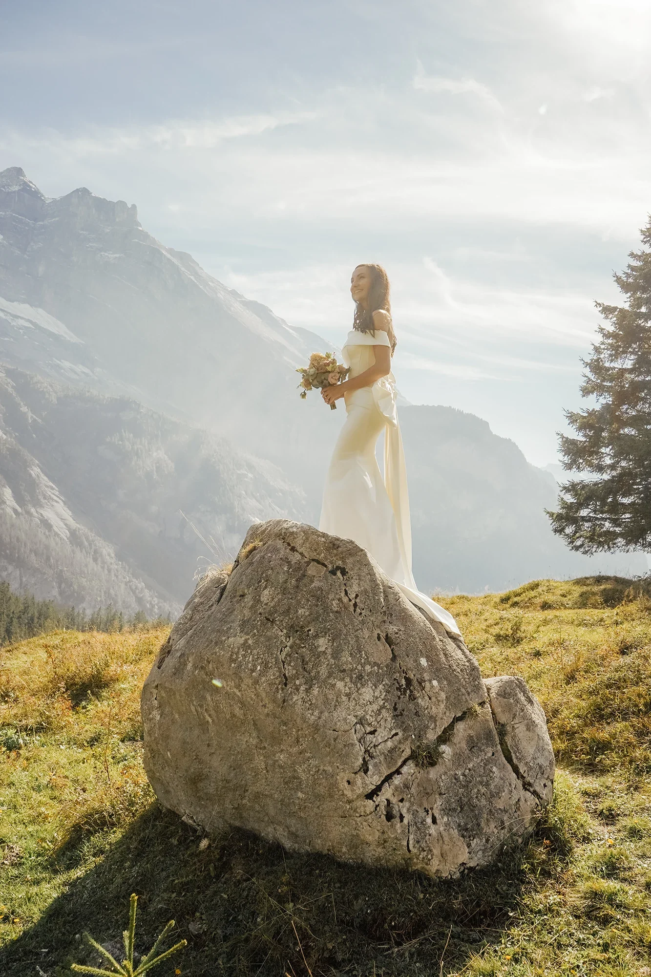 Eine Braut steht auf einem großen Felsen in einer Bergwelt mit schneebedeckten Gipfeln und Tannenbäumen im Hintergrund, während die Sonne scheint.