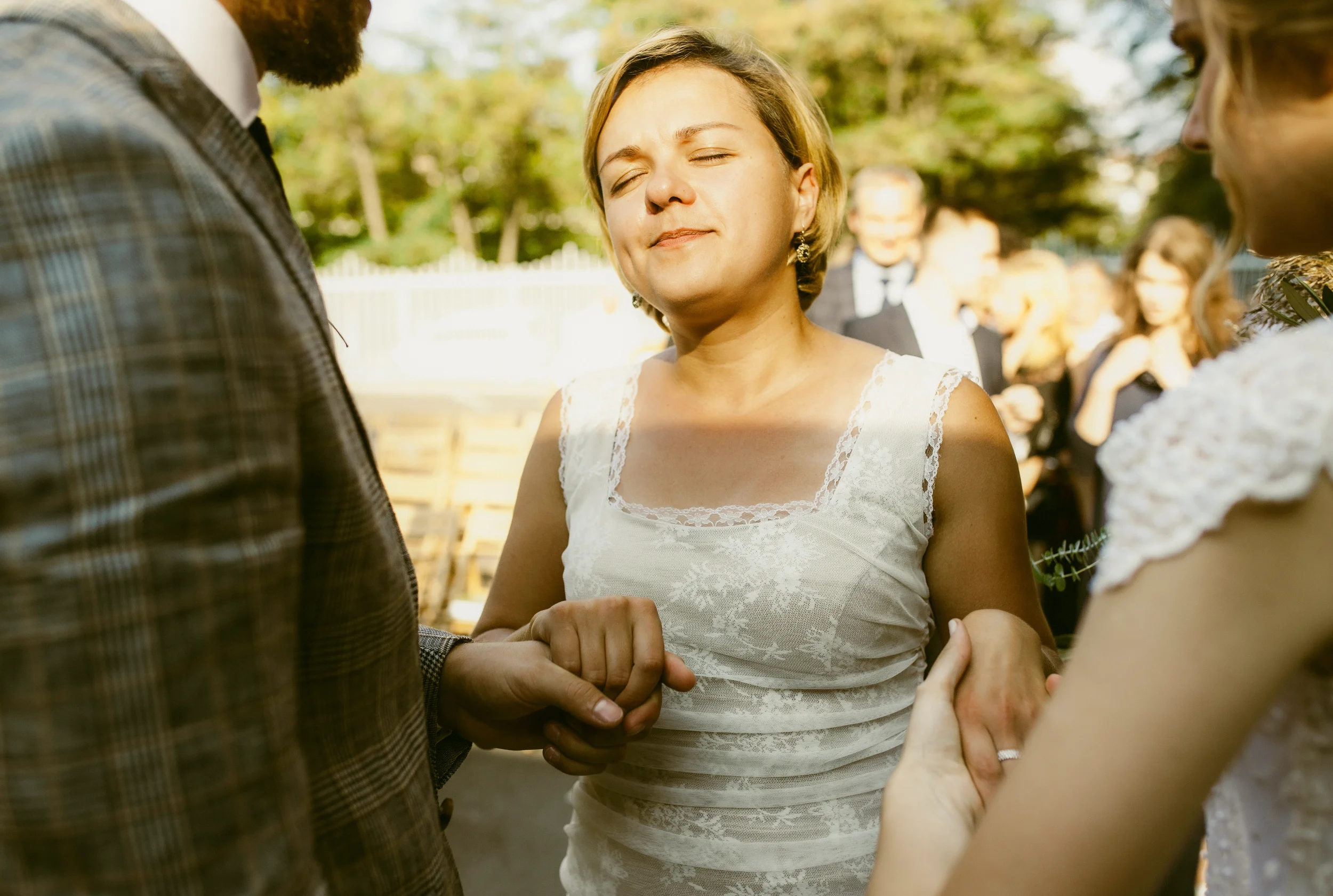 Eine junge Frau, wahrscheinlich bei einer Hochzeit, die einen feierlichen Moment im Freien erlebt. Sie ist umgeben von anderen Menschen, die offenbar ebenfalls an einem besonderen Anlass teilnehmen.