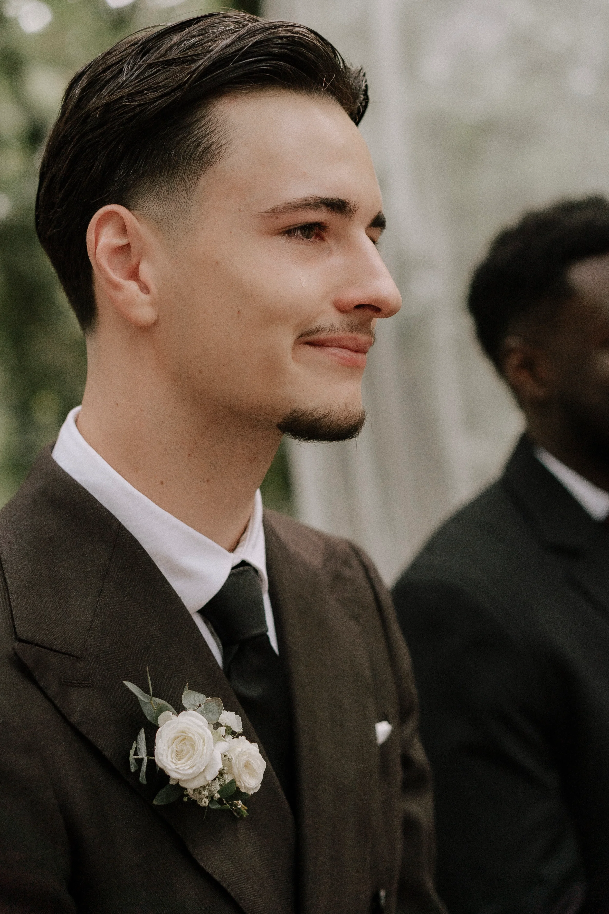 Nahaufnahme von einem Mann in formeller Kleidung mit Blumenbouquet auf der Brust, wahrscheinlich bei einer Hochzeit, mit Freunden im Hintergrund.