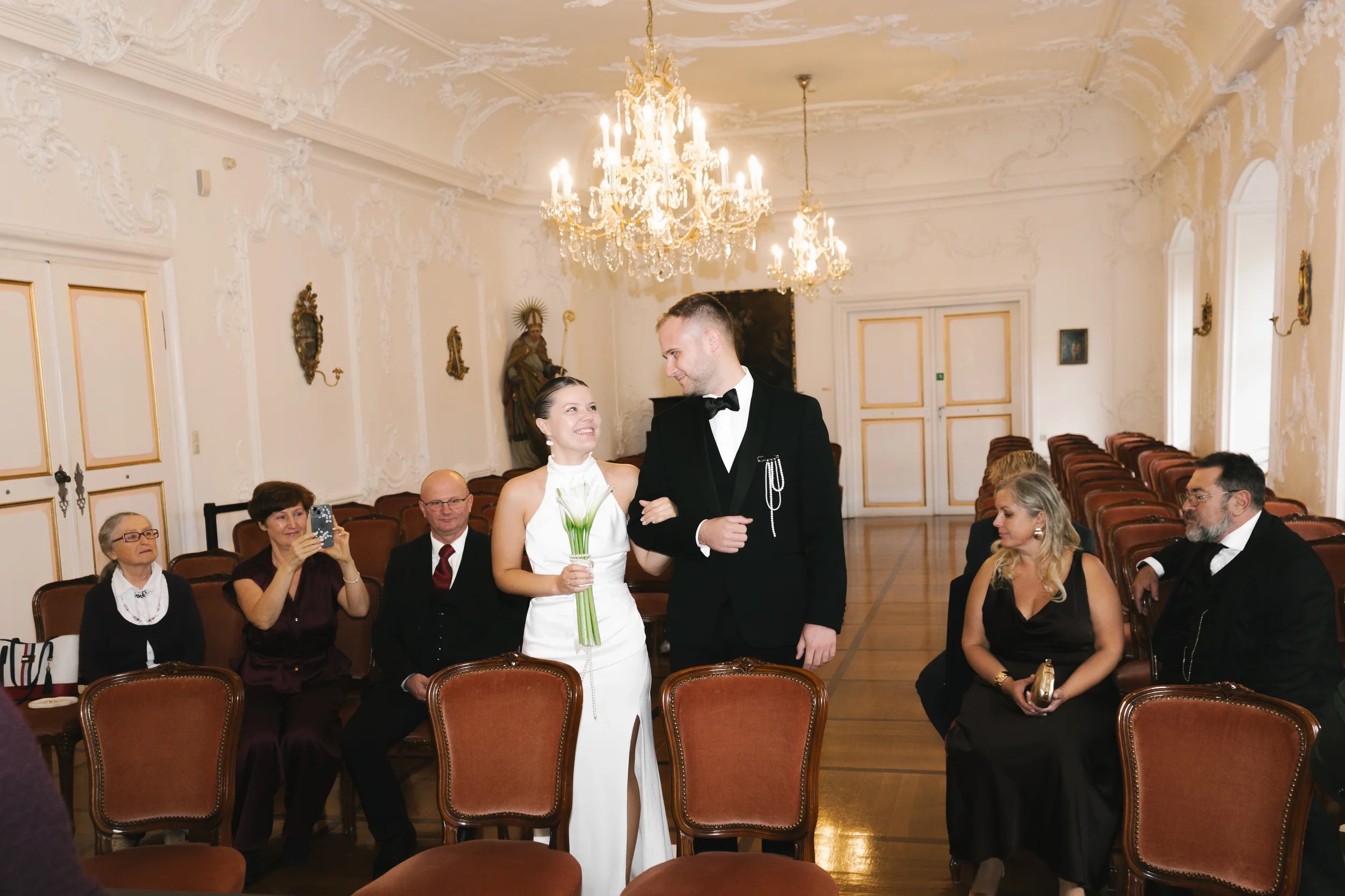 Ein Brautpaar steht in einem eleganten Saal vor einer Gruppe von Gästen. Die Braut trägt ein weißes Brautkleid und hält einen Blumenstrauß, während der Bräutigam einen schwarzen Anzug mit Fliege trägt. Die Gäste sitzen auf samtbezogenen Stühlen, eini