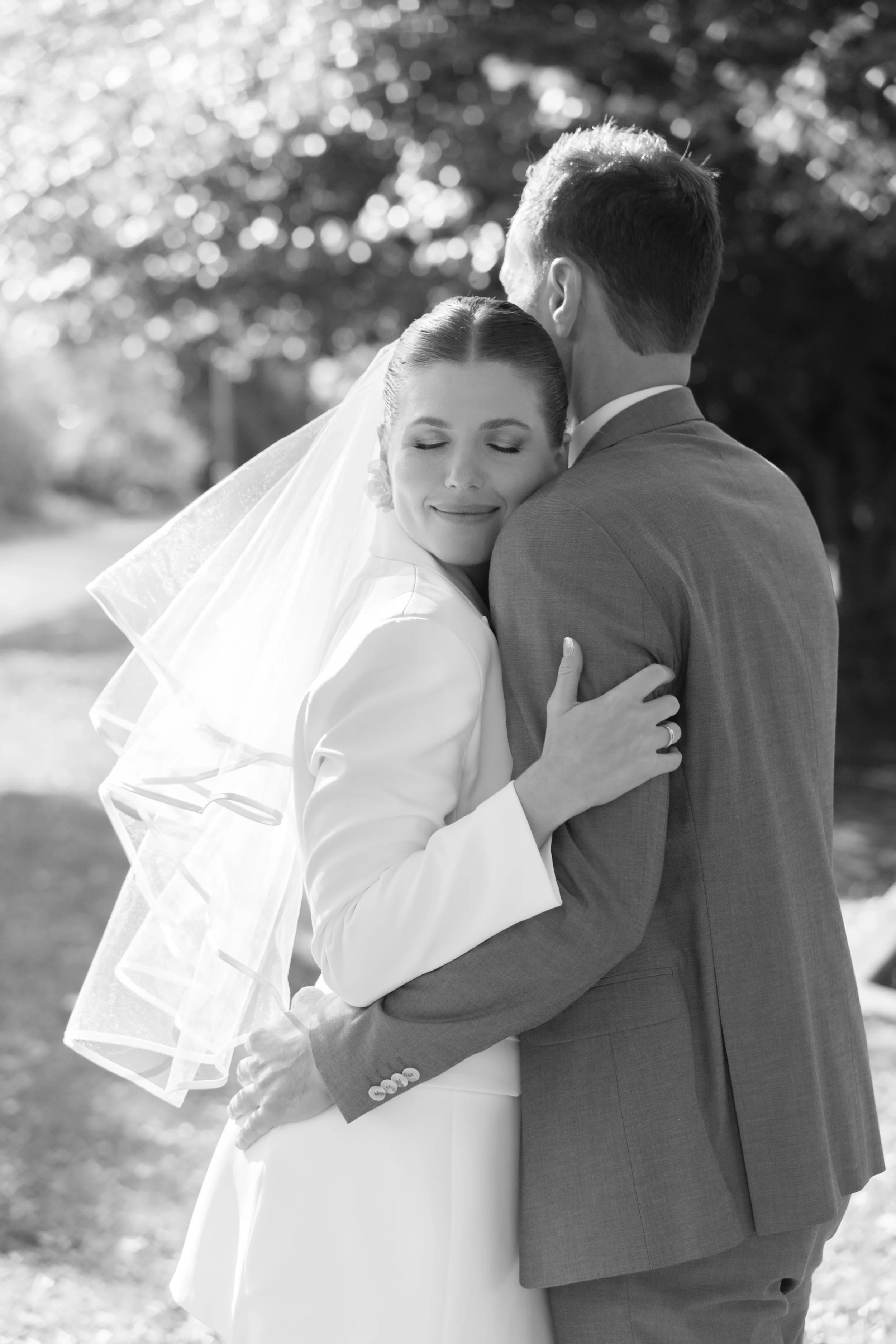 Schwarz-Weiß-Foto eines glücklichen Paares bei der Hochzeitsumarmung im Freien, mit Bäumen im Hintergrund.