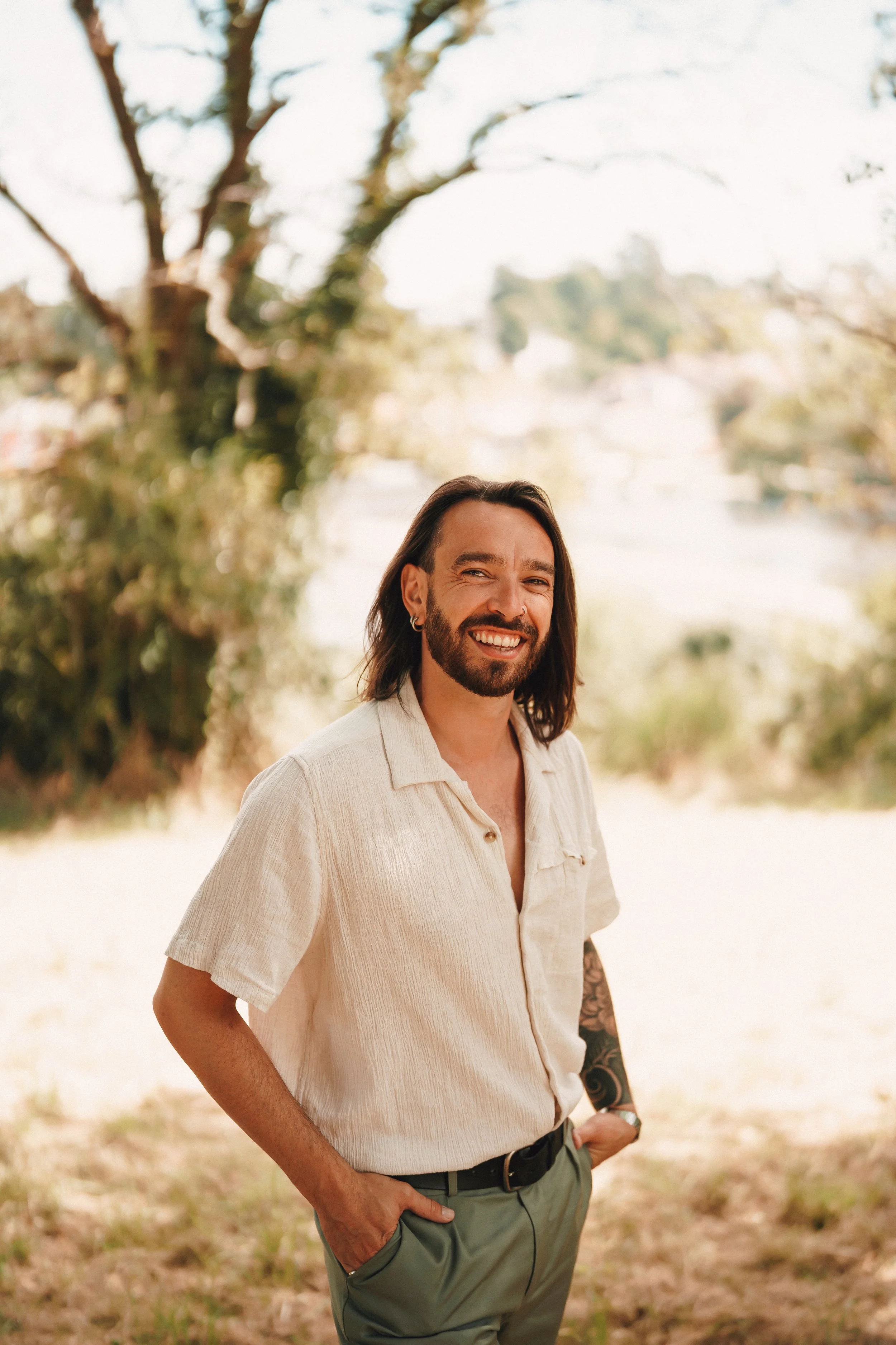 Un homme souriant, avec des cheveux longs et une barbe, portant une chemise beige à manches courtes et un pantalon vert, debout dans un environnement extérieur avec des arbres en arrière-plan.