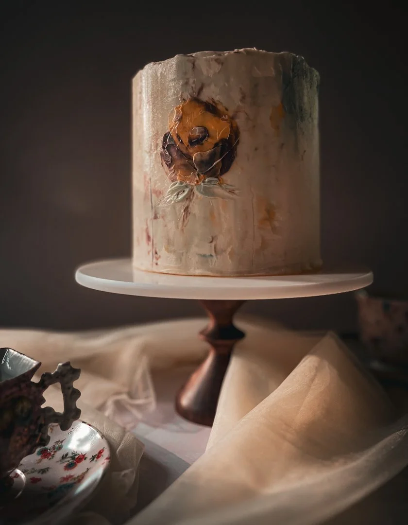 Tall neutral cake with abstract floral painting in warm ochre tones on a white cake stand, Grand Rapids, MI