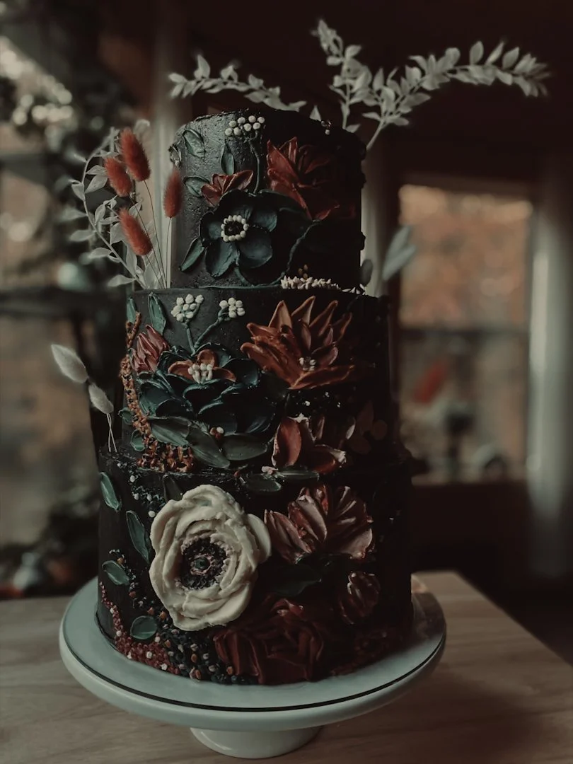 Moody charcoal wedding cake with sculpted florals, foliage, and deep rust accents on a white cake stand, Grand Rapids, MI