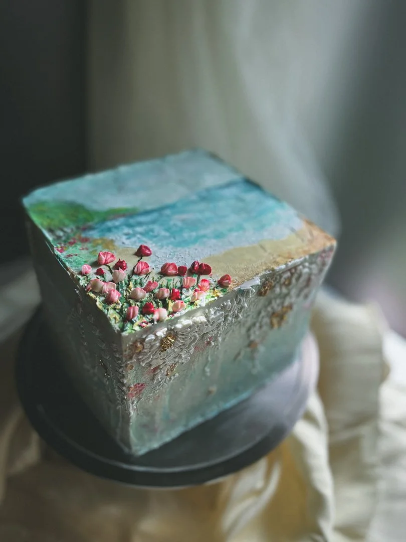 Custom cake designed to look like a painting of a beach with pink textured flowers place at the bottom corner of the cake.