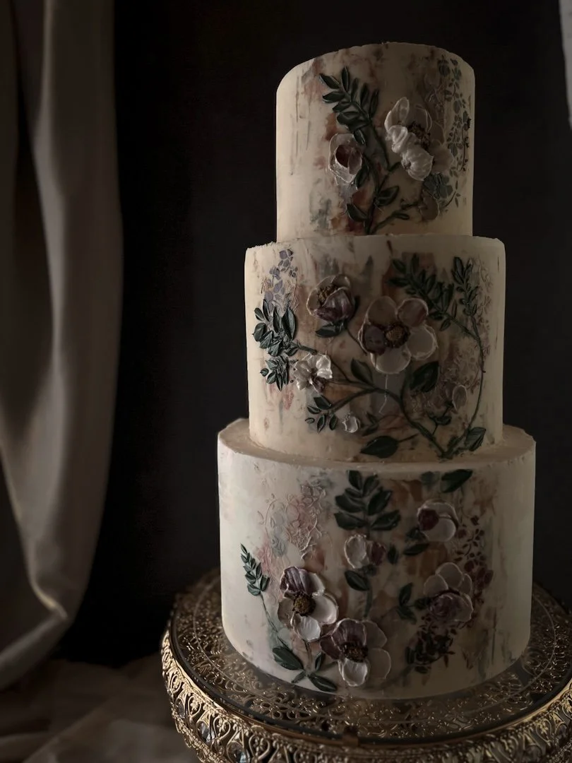 Three-tier ivory wedding cake with European-inspired floral and leaf artwork on an ornate gold cake stand