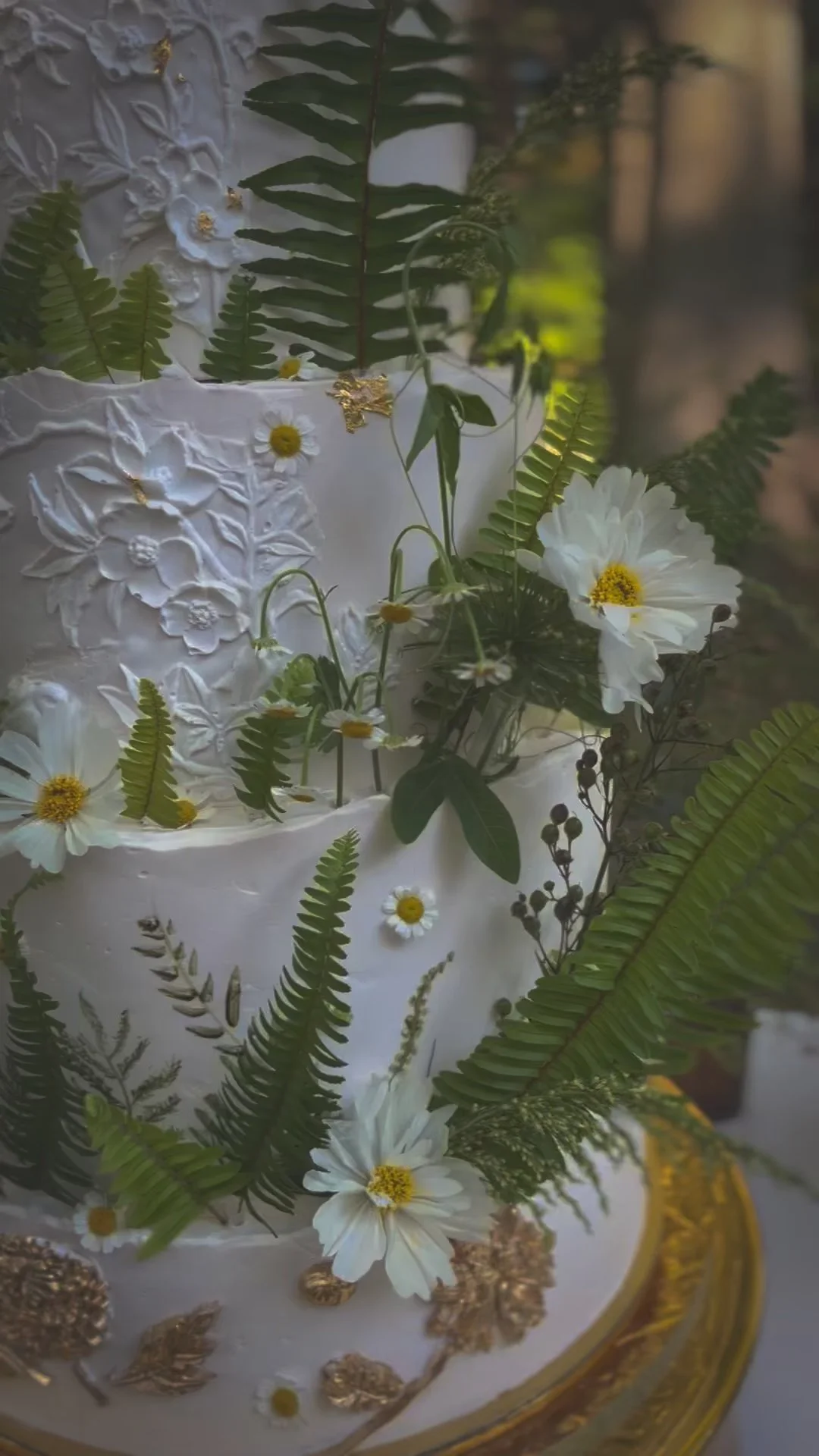 Close-up of a white decorated cake adorned with white flowers, daisies, fern leaves, and gold accents, set on a gold-rimmed cake stand.