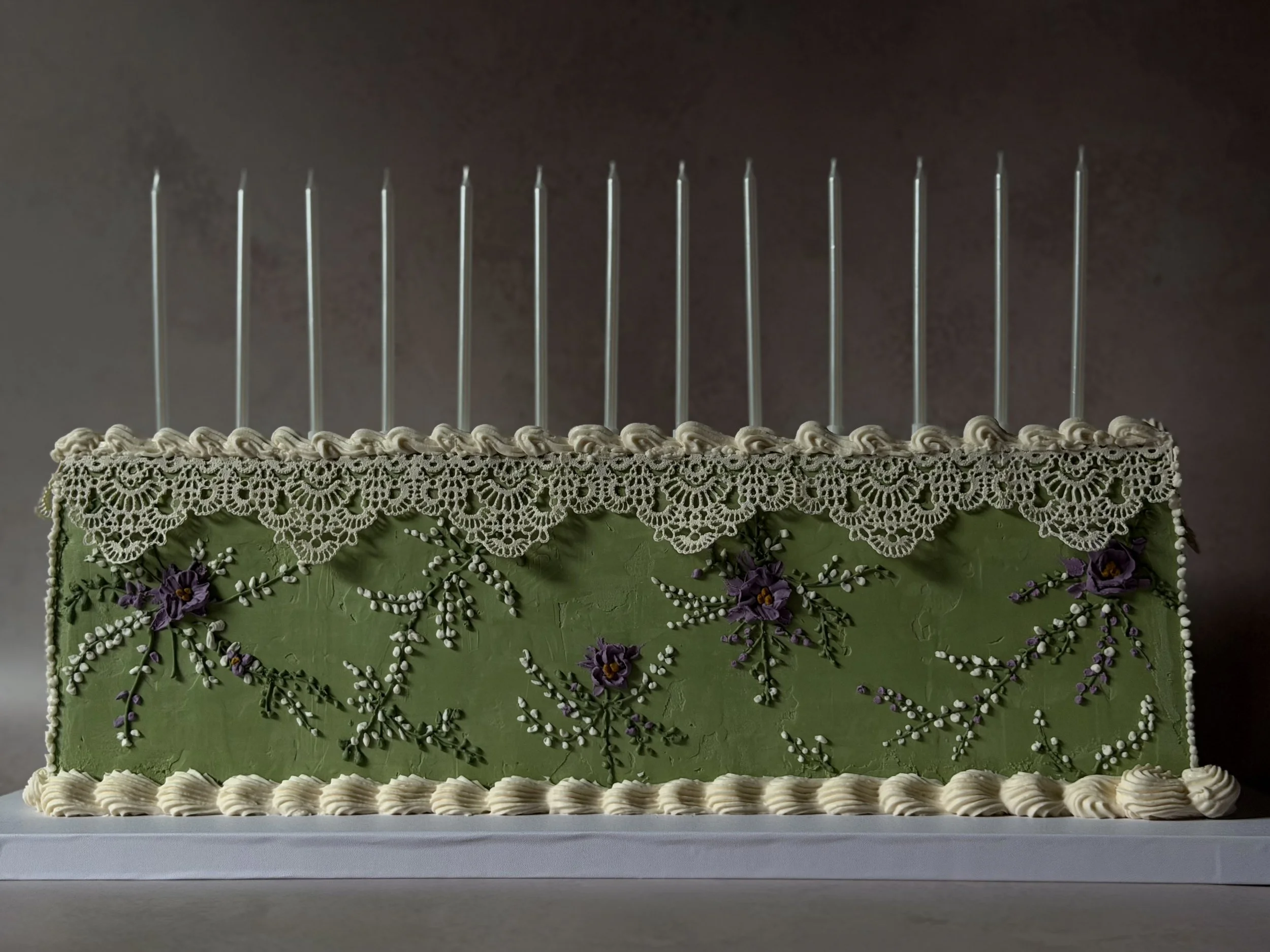 Vintage-inspired green rectangular cake with piped borders, lace trim, hand-painted florals, and tall candles