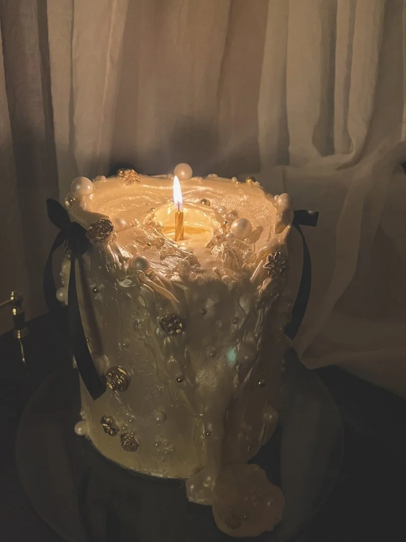 White candle cake with flickering flame, pearl details, and gold accents on a dark surface with draped fabric