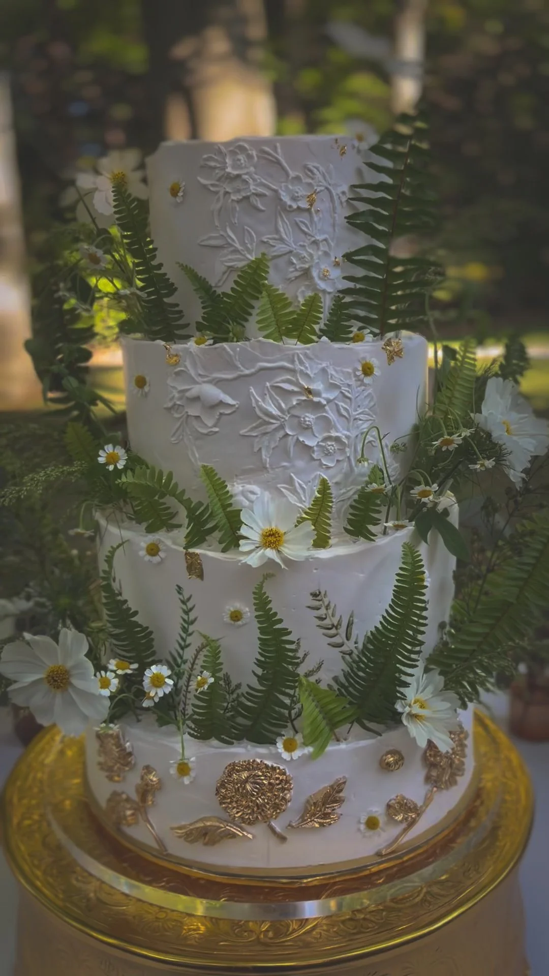 A white four tier wedding cake with buttercream floral textures, fresh ferns, daisies, cosmos, and gold leaf accents.