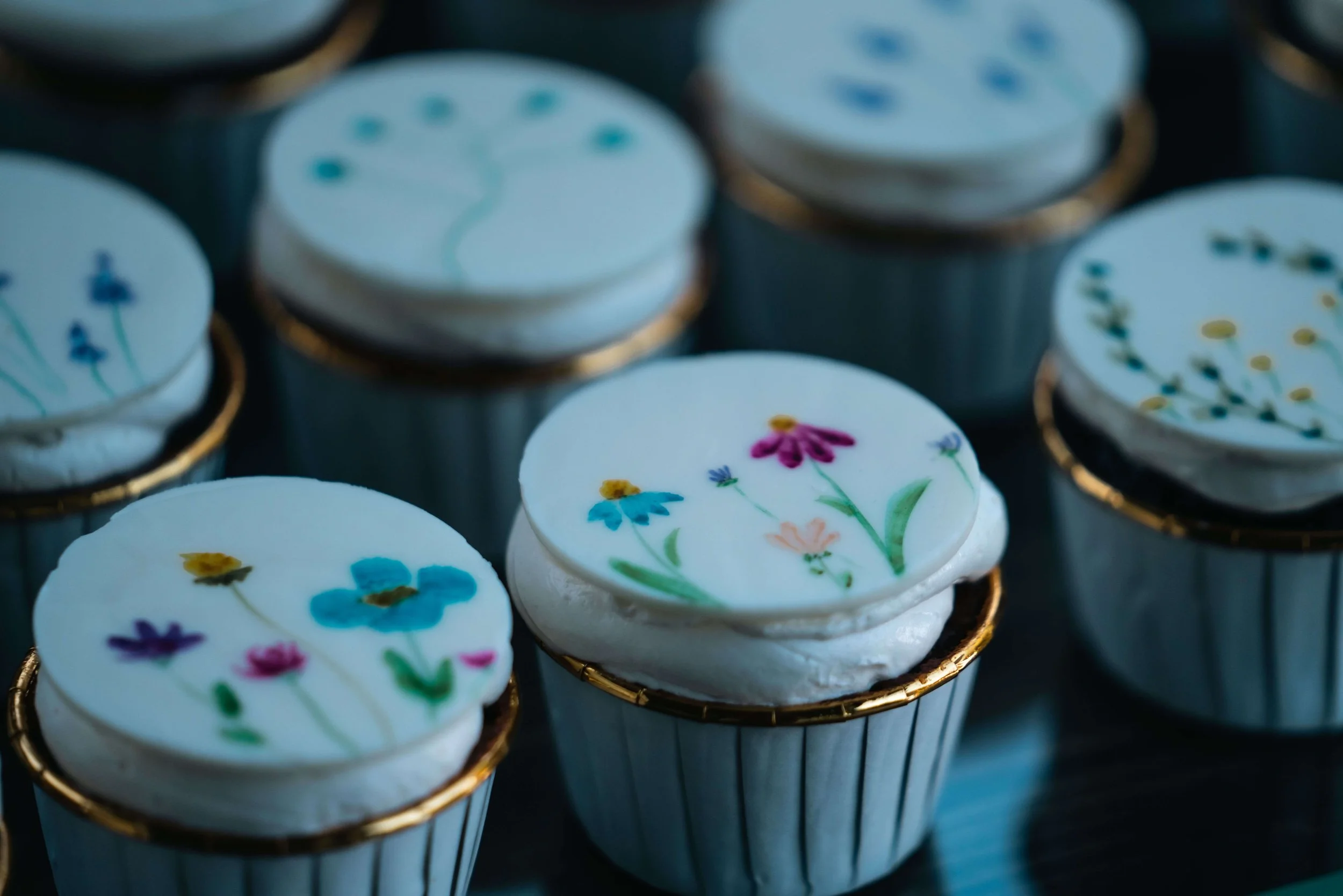 Cupcakes with hand-painted wildflower designs on white chocolate tops, styled with gold-trim wrappers, West Michigan