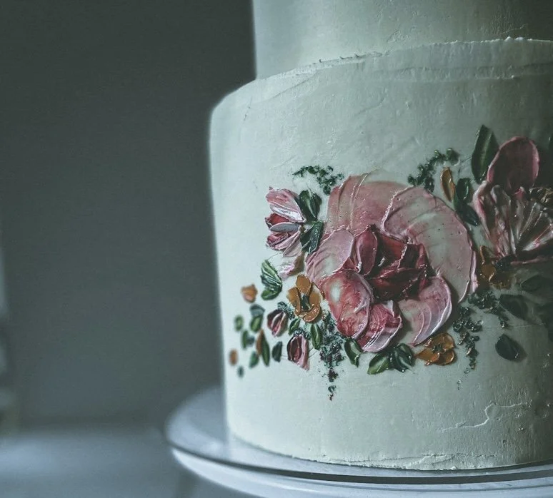Close-up of a white cake decorated by Artfully Meraki, with hand-painted pink and red flowers and green leaves.