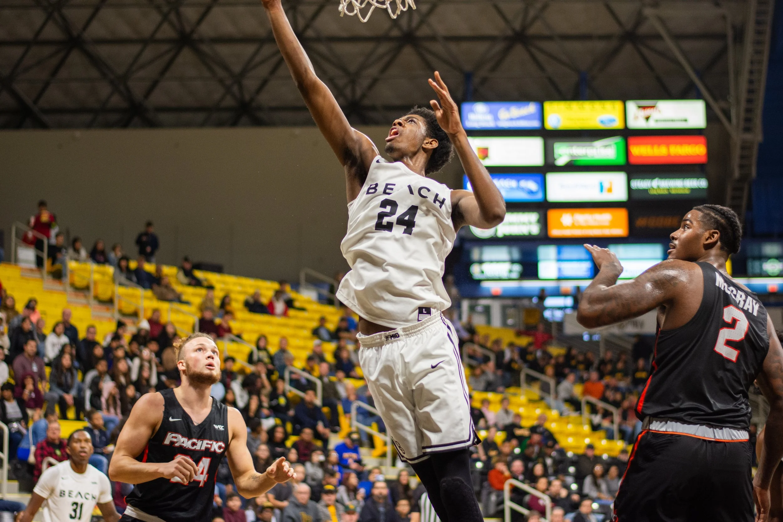 A basketball player in a white jersey with the number 24 jumps to make a shot or block a shot against a player in a black jersey with the number 2. The game is taking place in an indoor basketball court with spectators in the background and several a