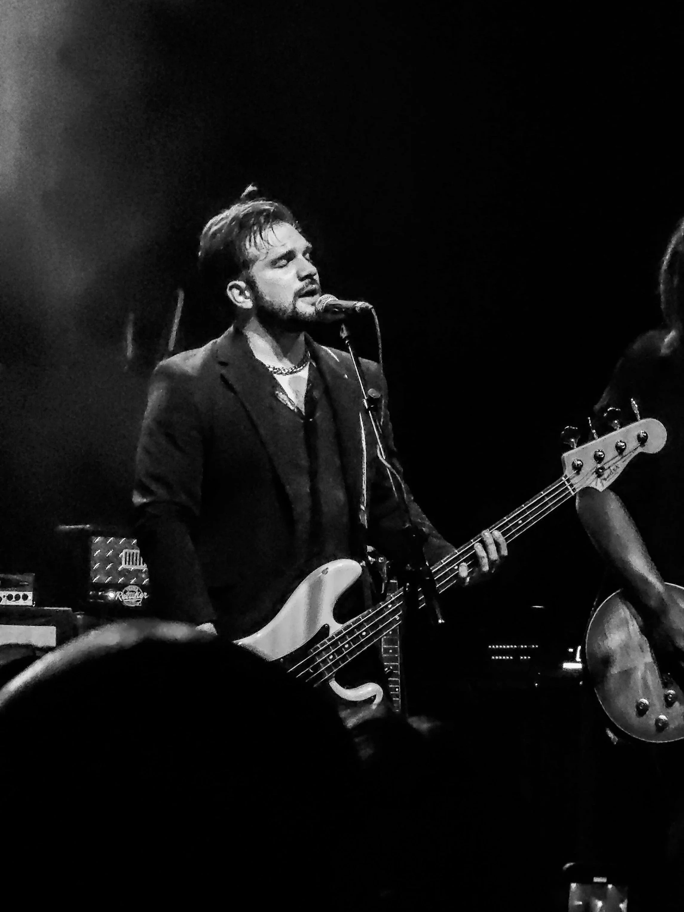 A black-and-white photo of the bass play of the band Classless Act singing into a microphone while playing an electric guitar on stage, with musical equipment in the background.