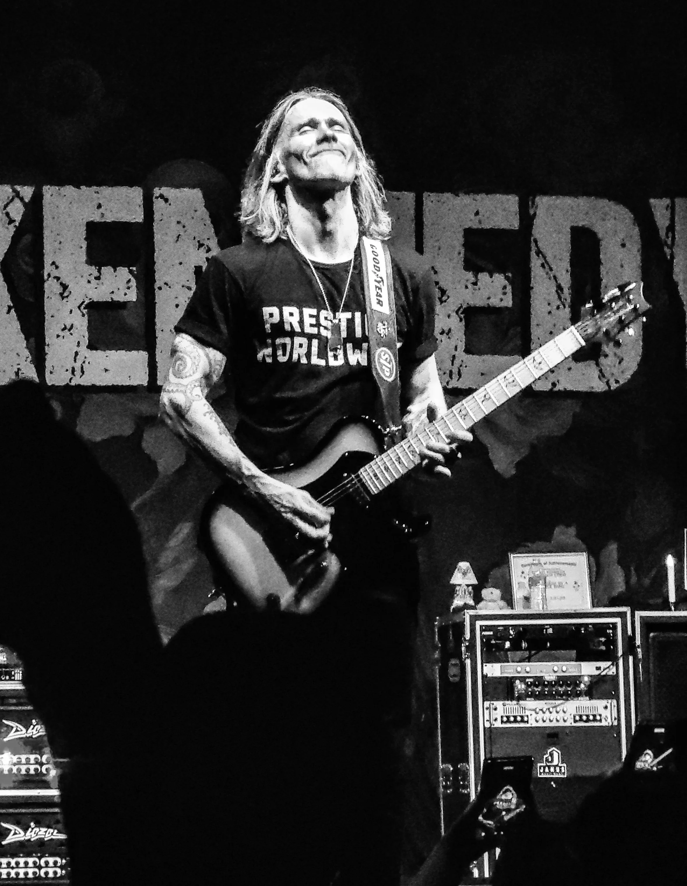 A black and white photo of Myles Kennedy performing on stage with a guitar, standing in front of a backdrop with the word "KENNEDY" in large, distressed letters.