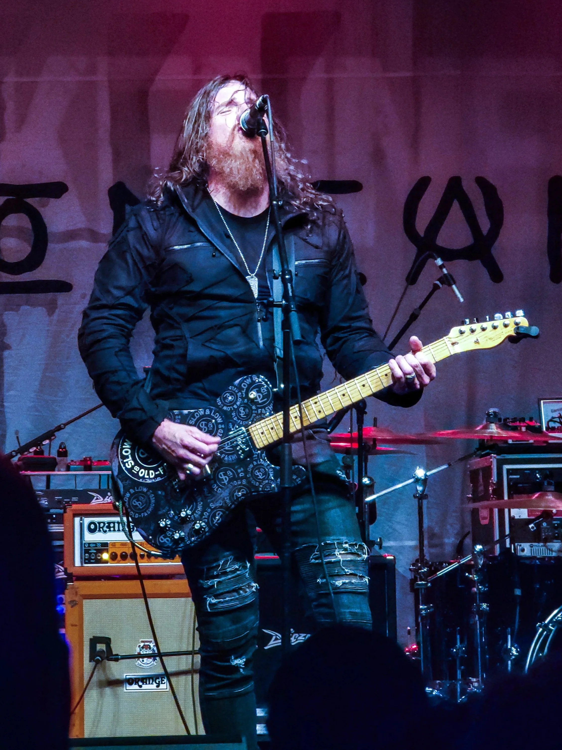 A male musician with long hair and a beard, wearing a black jacket and ripped jeans, is singing into a microphone while playing an electric guitar on stage. Musical equipment and amplifiers are visible in the background.