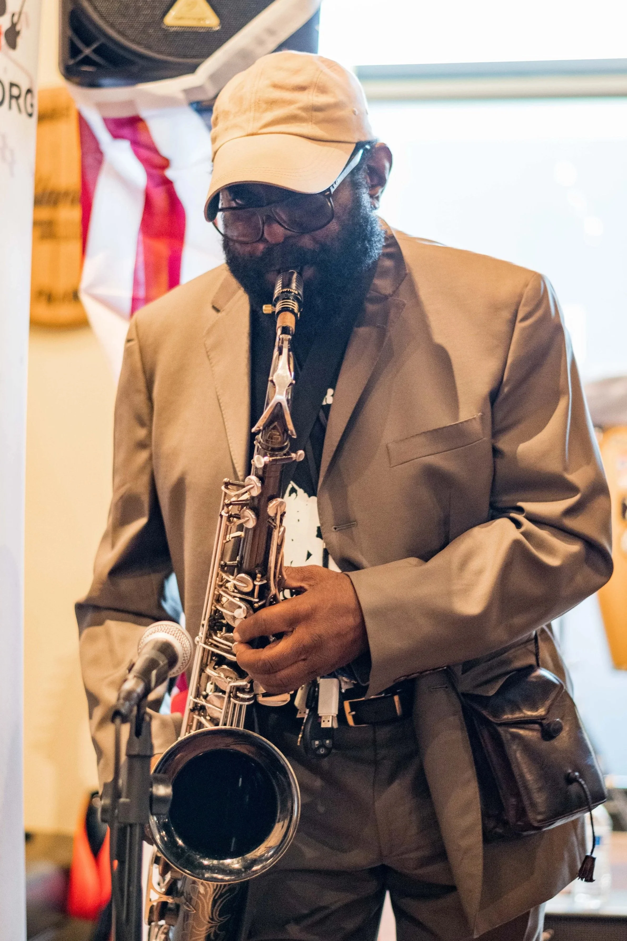 Man playing a saxophone at an event, wearing a beige hat, sunglasses, and a tan blazer, with a backdrop of a banner and equipment.
