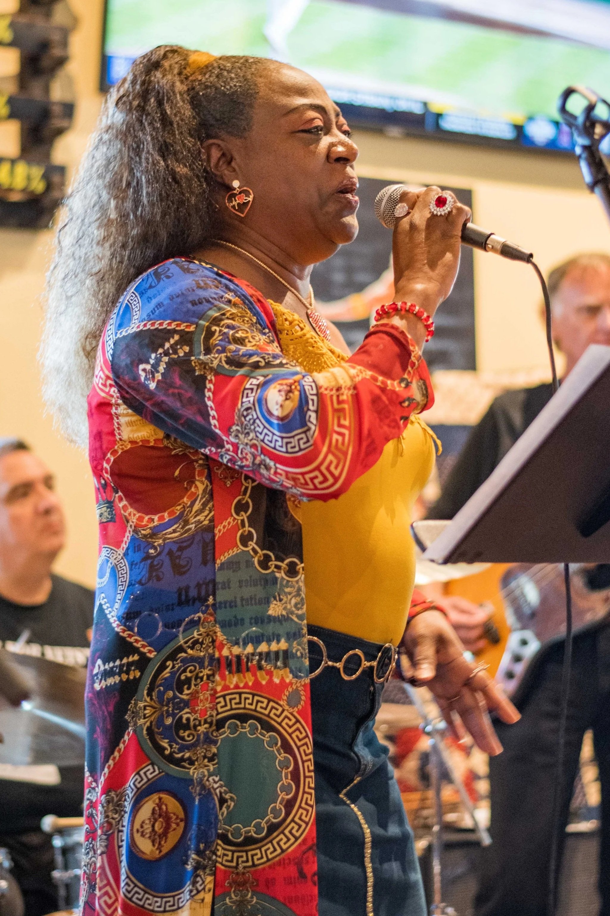 A woman singing into a microphone at an indoor event. She is wearing colorful, patterned clothing with gold accents, earrings, a necklace, and rings.