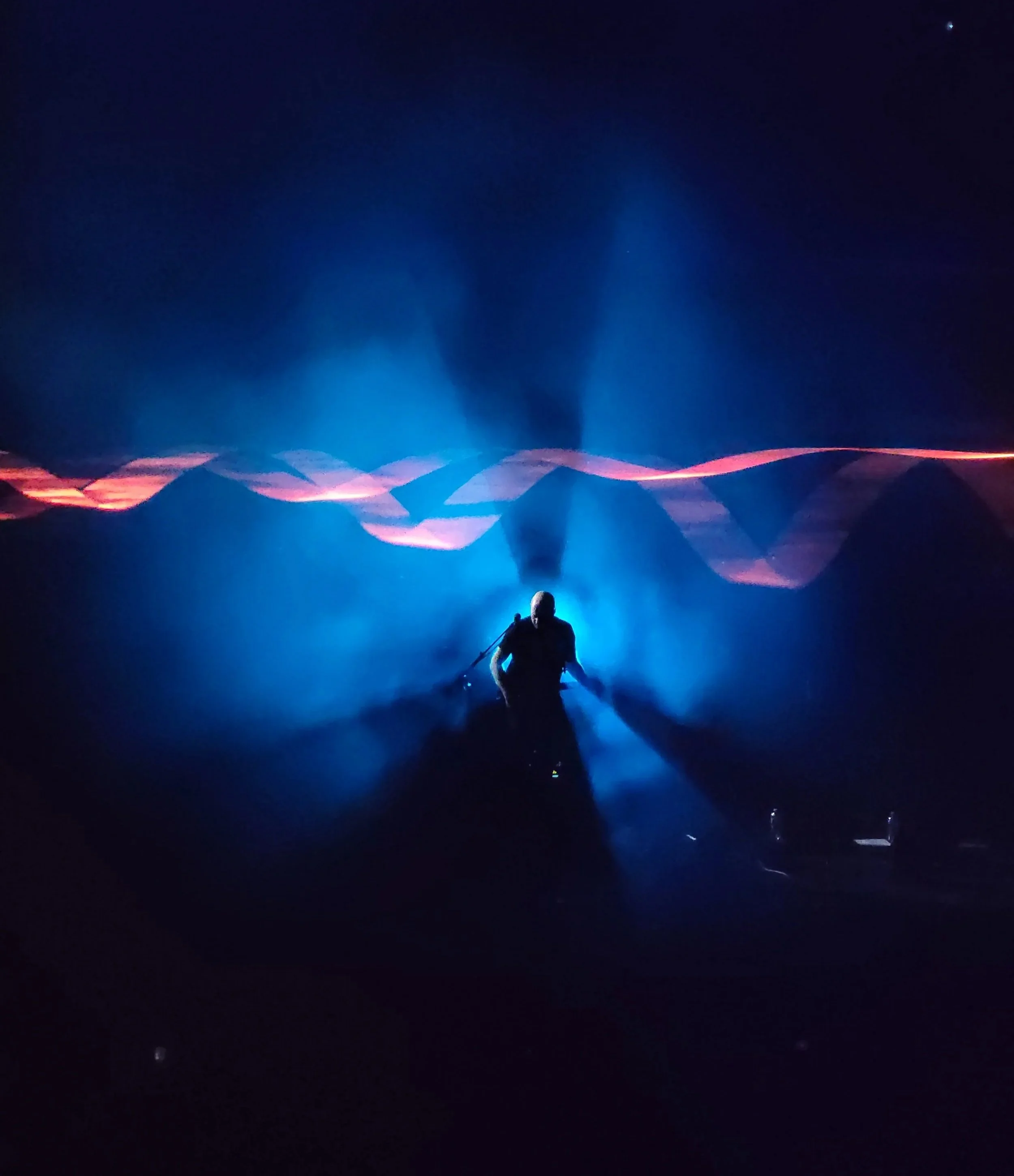 The guitarist of the band Brit Floyd standing on stage with blue lighting against a dark background with abstract light patterns.