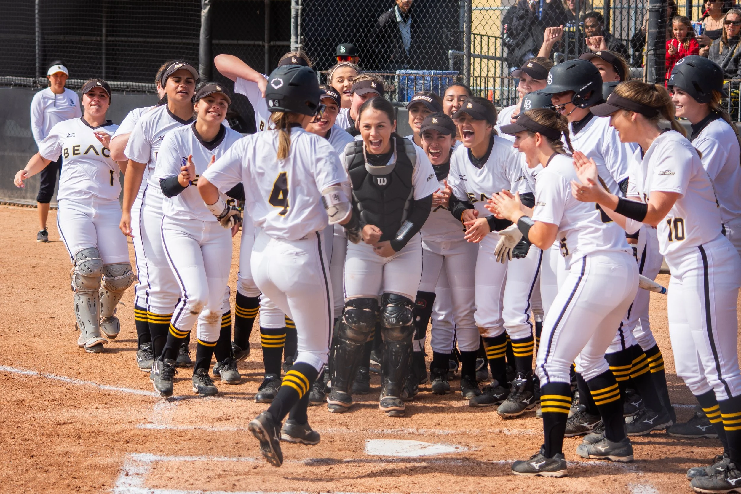 A group of female softball players celebrating on the field, with some wearing helmets and others with their hands raised, while fans watch in the background.