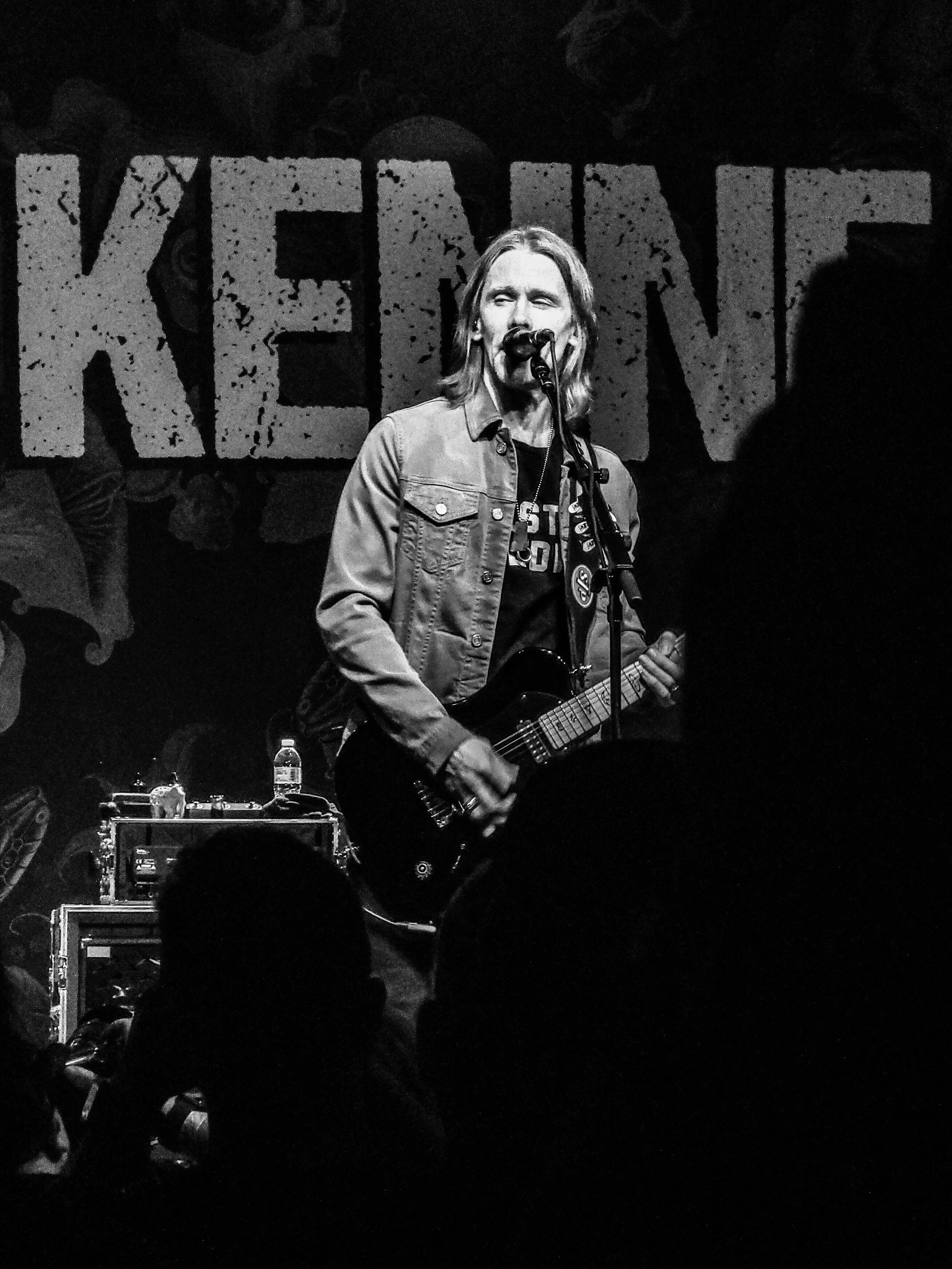 A black and white photo of Myles Kennedy performing on stage with a guitar, standing in front of a backdrop with the word "KENNEDY" in large, distressed letters.