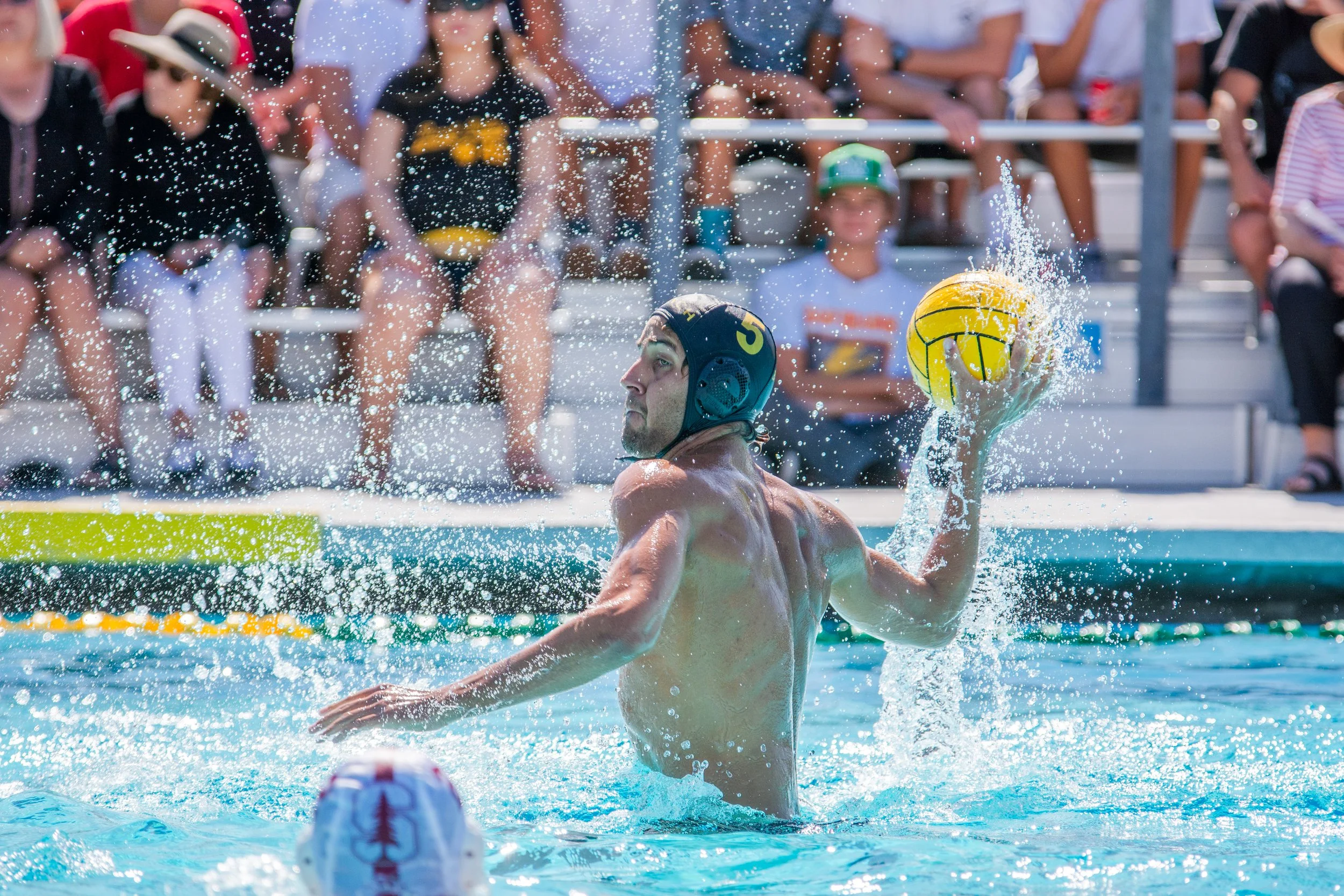 A water polo player wearing a black cap with the number 5, holding a yellow water polo ball, in the swimming pool with spectators in the background.