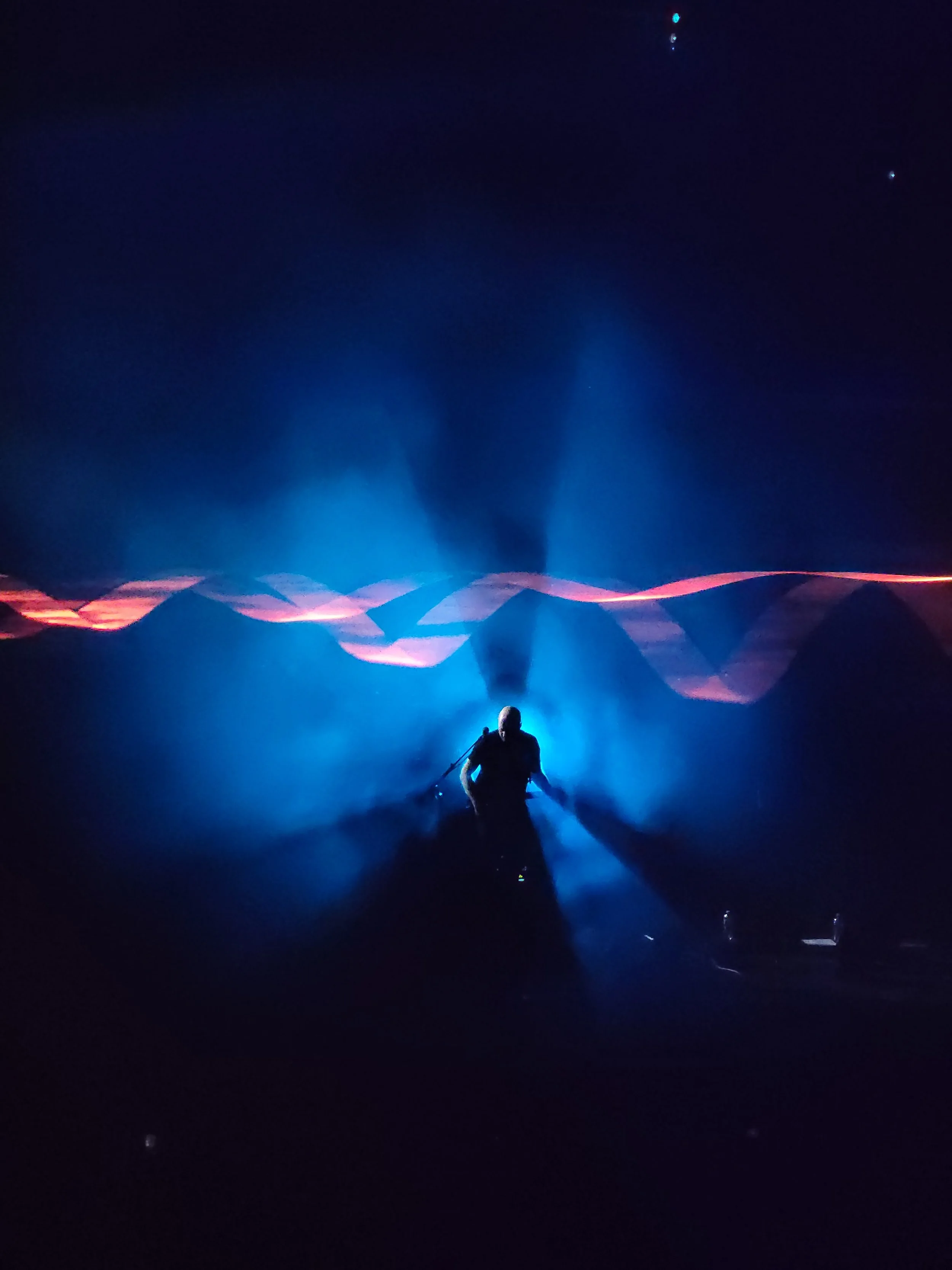 The guitarist of the band Brit Floyd standing on stage with blue lighting against a dark background with abstract light patterns.