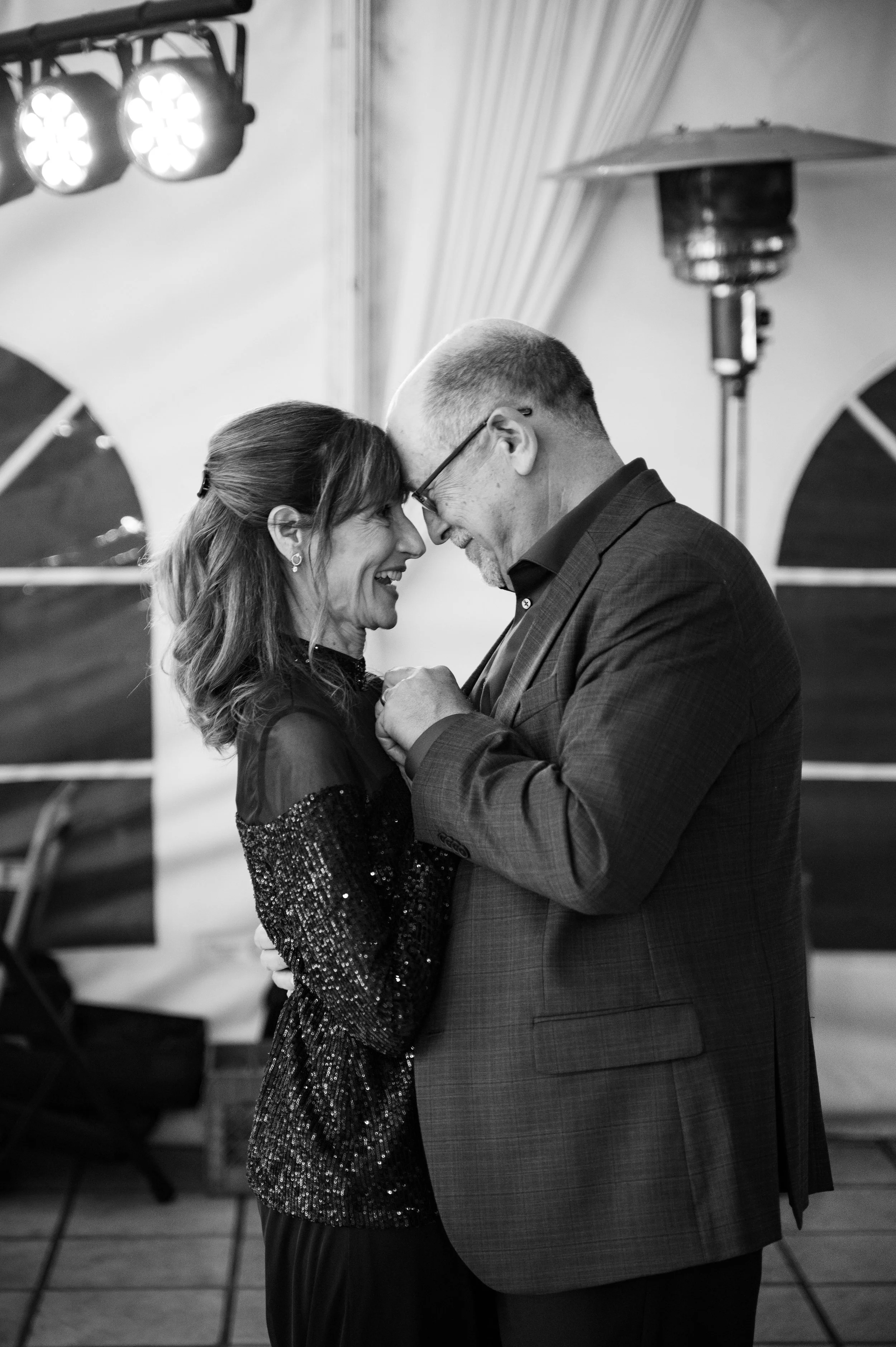 A black-and-white photo of a couple sharing a dance, with their foreheads touching and smiling at each other. The woman is wearing a sparkly dress and has her hair down, while the man is wearing a suit and glasses. They are close together, holding ha