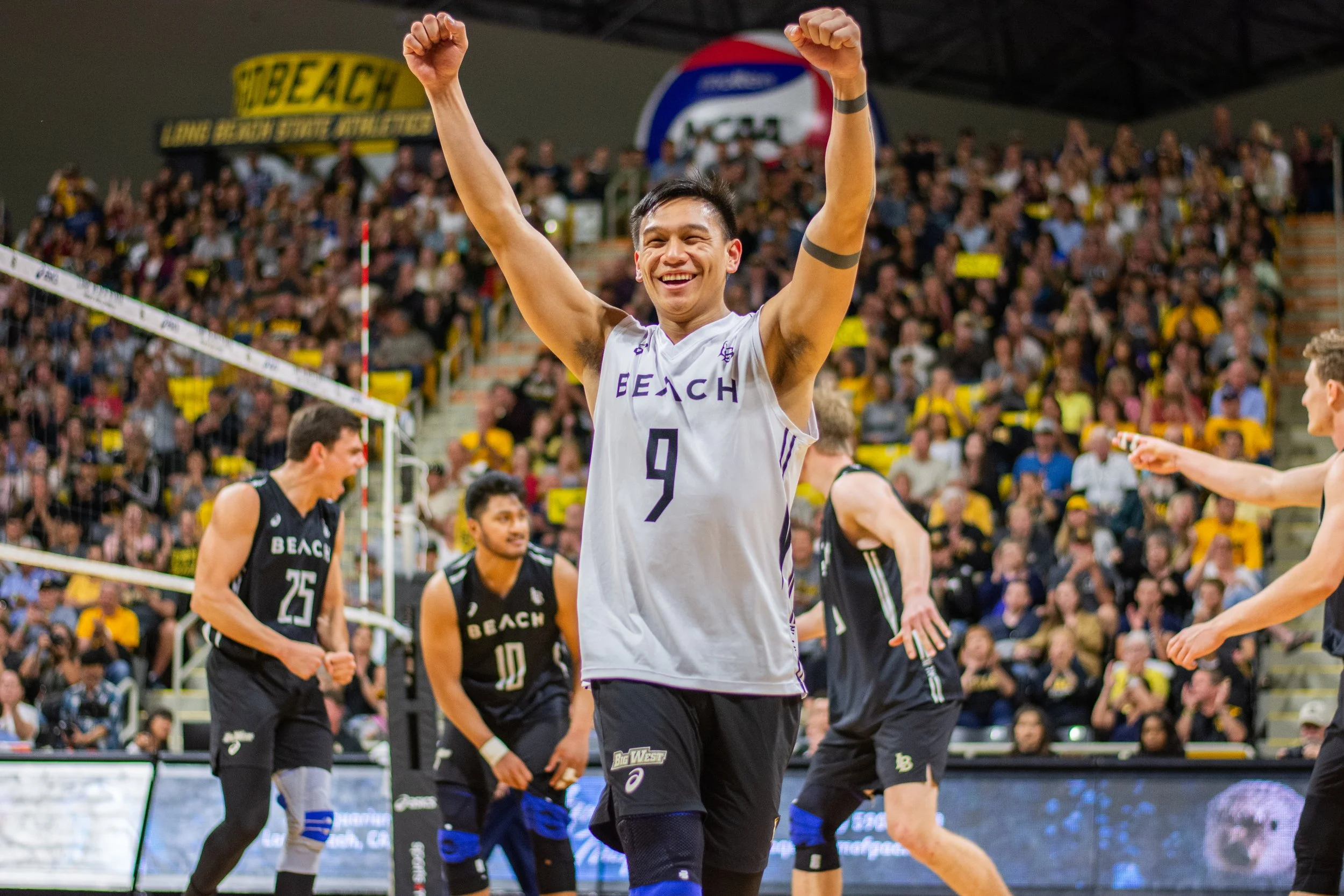 A volleyball player wearing a white jersey with the number 4 and the word BEACH on it celebrates with arms raised in front of a large crowd at an indoor volleyball court.