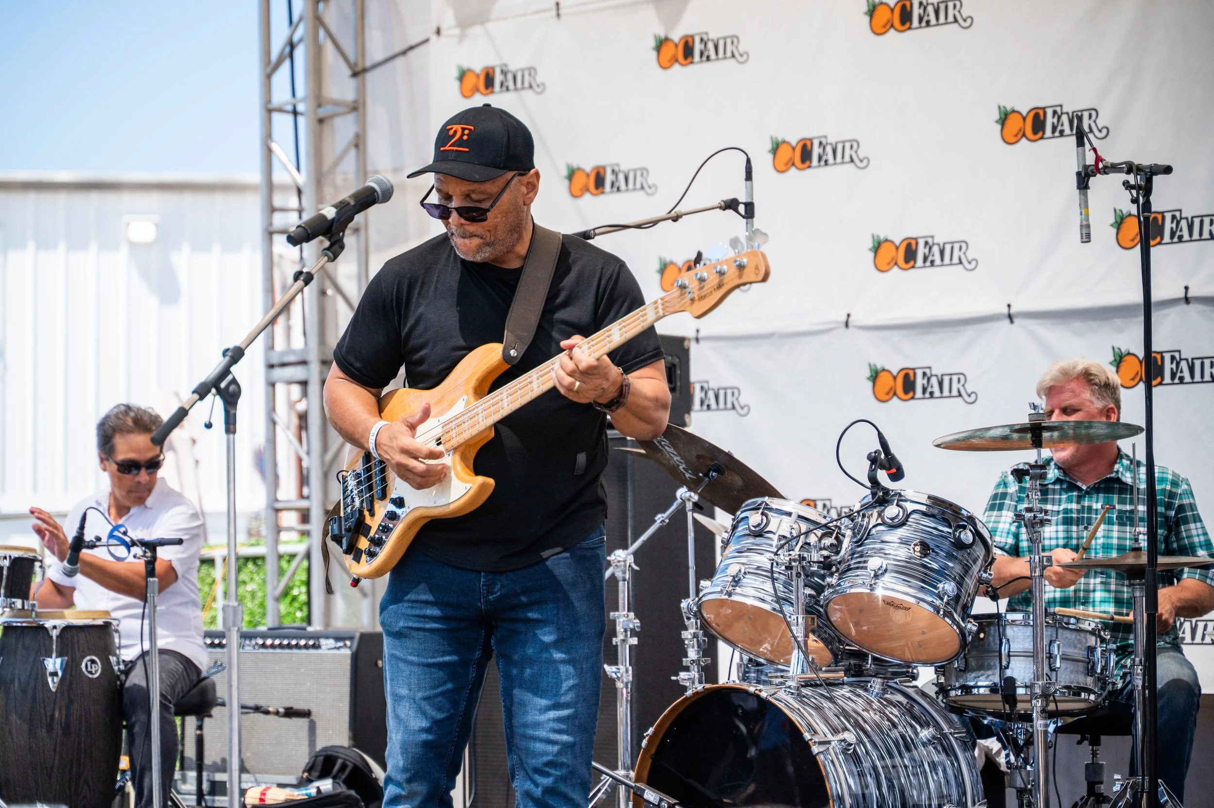 A group of musicians performing live on an outdoor stage with OC Fair banners in the background. The image features a man playing an electric bass guitar, a drummer, and other band members.