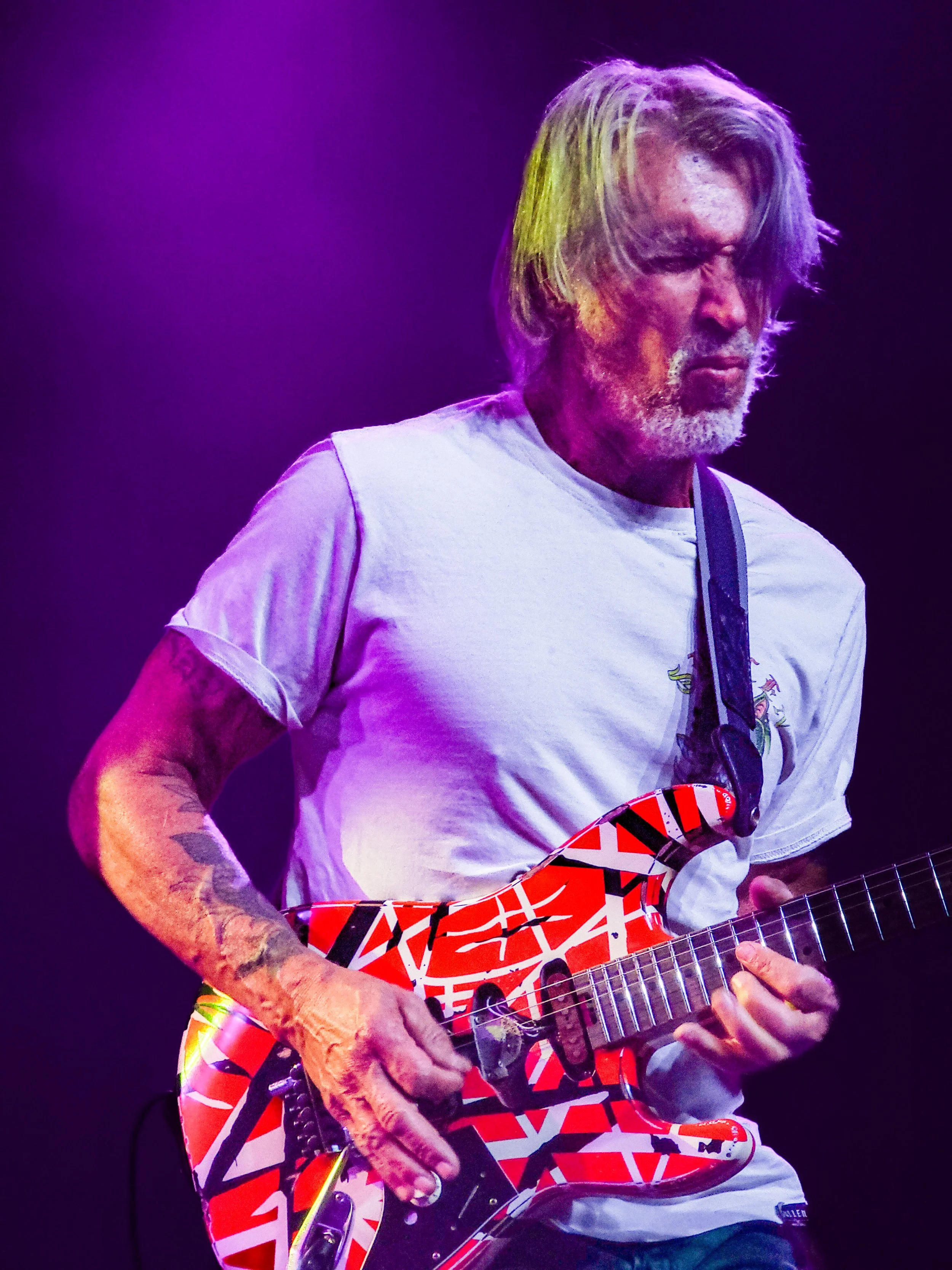 George Lynch playing an electric guitar with a red, white, and black geometric pattern on the body during a concert. The stage is lit with purple lighting.