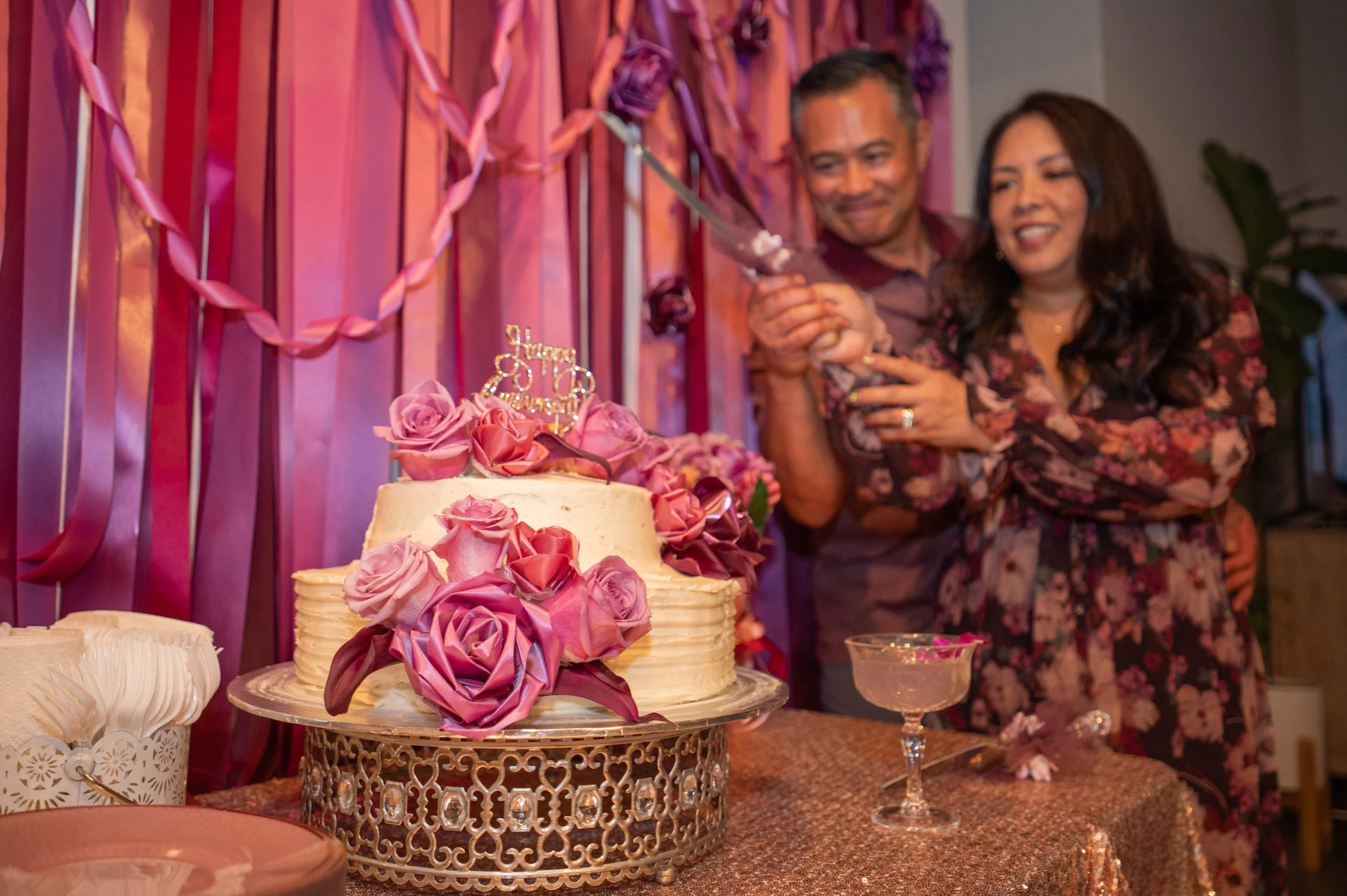 A wedding cake decorated with pink and purple roses on a golden cake stand. Two people are in the background, smiling and holding a dessert serving utensil, with pink ribbon and balloon decorations on the wall behind them.