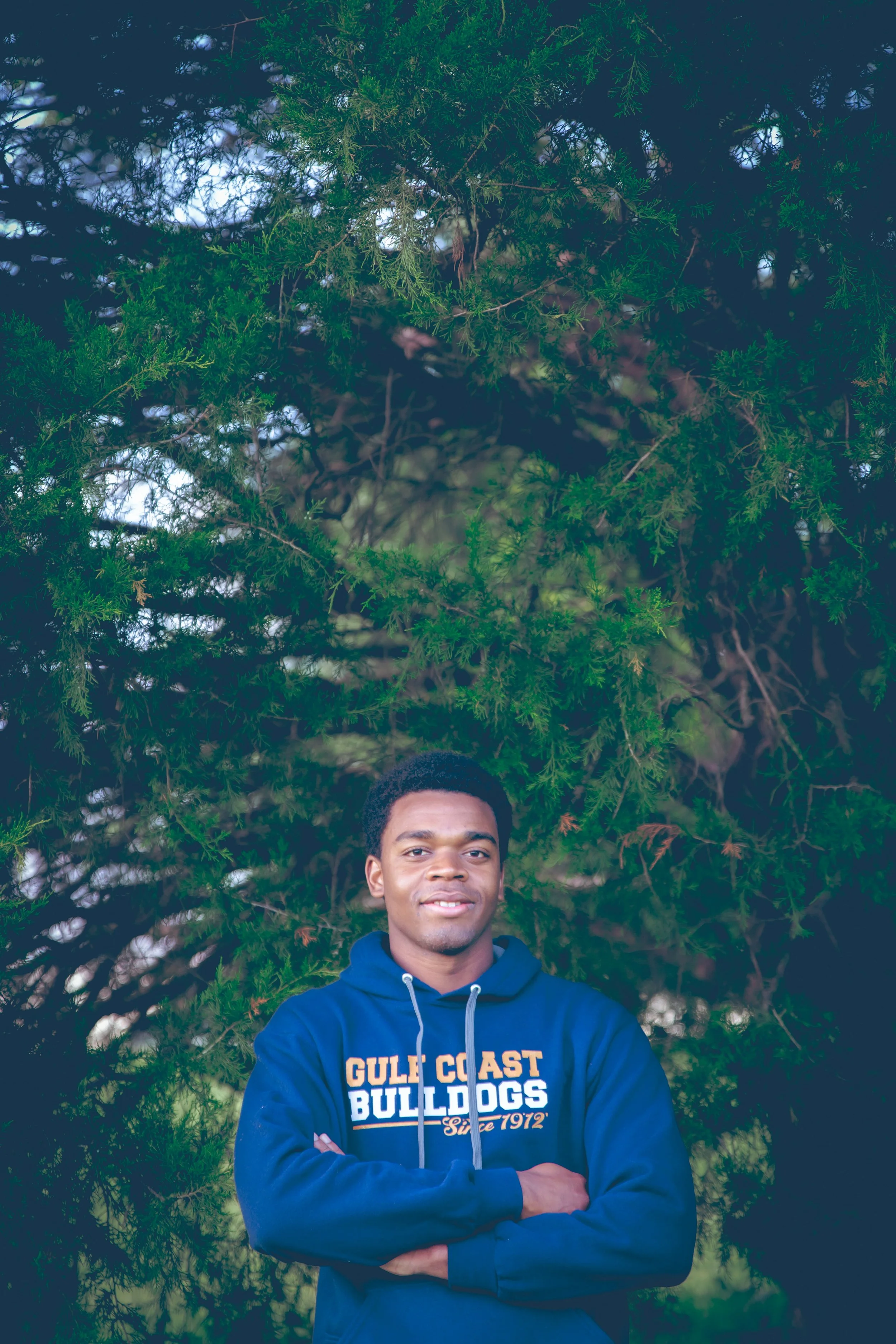 Young man standing outdoors in front of dense green pine trees, wearing a navy blue hoodie with 'Gulf Coast Bulldogs Since 1972' written on it, arms crossed, looking at the camera.  Senior Photographer Madison, MS 